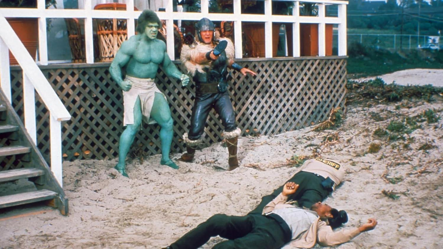Der Hulk und ein Krieger stehen sich gegenüber, während ein Sheriff im Sand liegt. Im Hintergrund sind eine Holzkonstruktion und Treppen zu sehen.