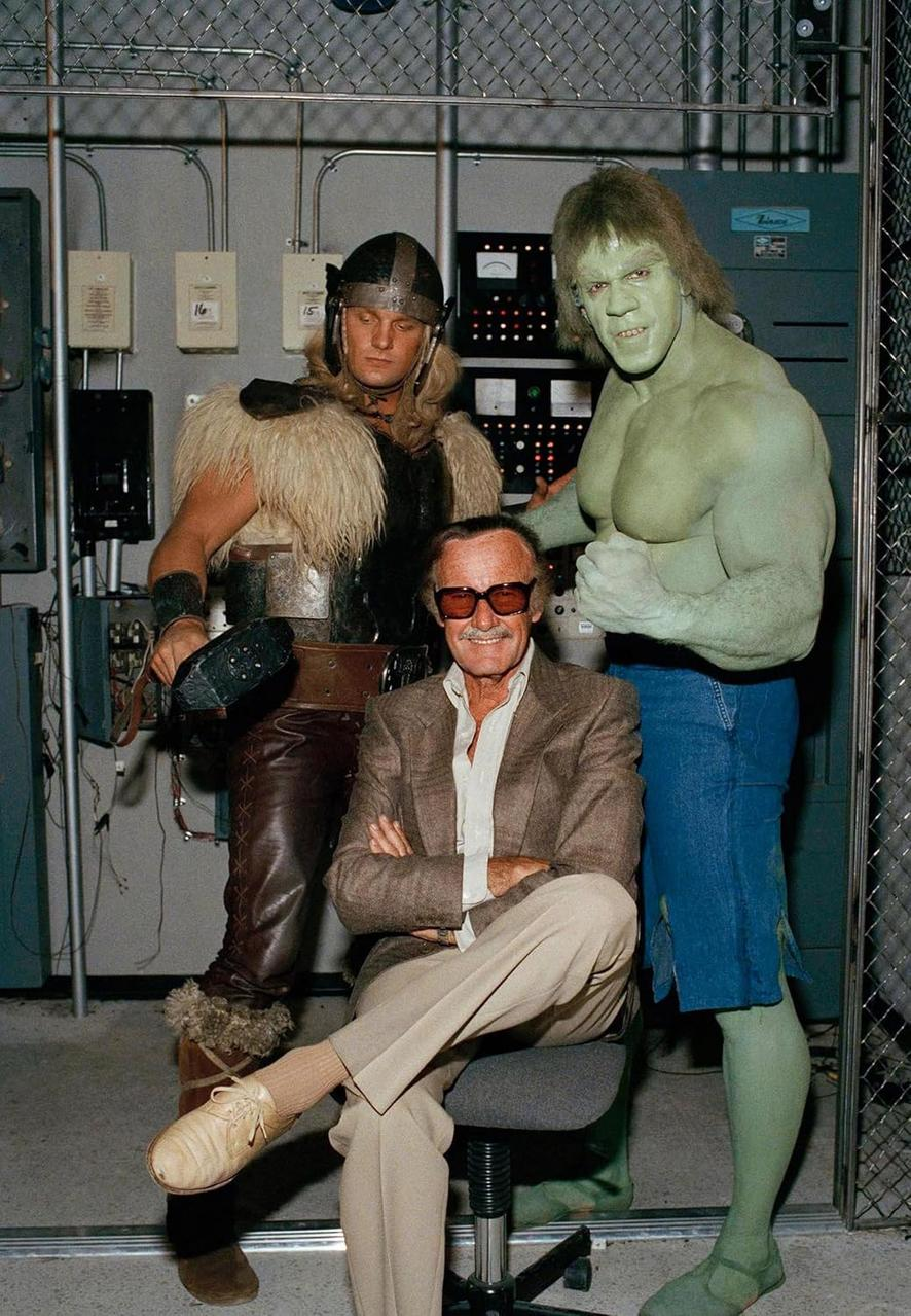 Stan Lee mit Hulk- und Thor-Darstellern. Lee sitzt, andere stehen. Industrielle Kulisse mit Ausrüstung und einem Maschendrahtzaun.