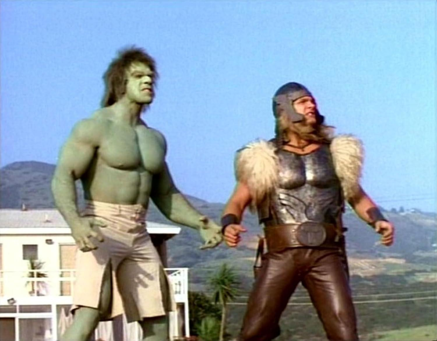 Der Hulk, grüne Haut, und Thor, in Rüstung, stehen zusammen. Im Hintergrund sind ein Haus und eine Landschaft unter blauem Himmel zu sehen.