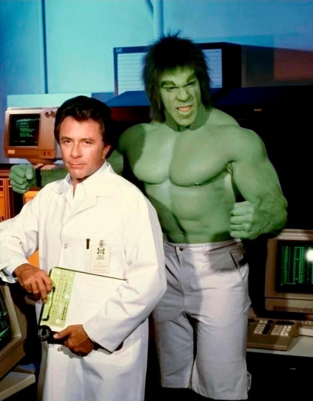 Der Hulk steht neben einem Mann im Laborkittel. Der Hulk ist grün und wütend. Der Hintergrund hat Computer und eine Laborumgebung.
