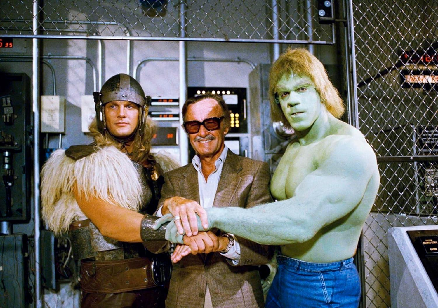 Stan Lee mit Thor- und Hulk-Darstellern, alle halten Hände. Metallkäfig und Geräte im Hintergrund.