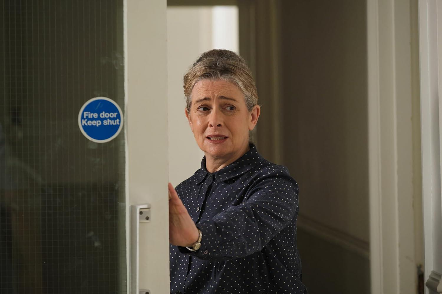 Eine Frau im Polka-Dot-Hemd öffnet eine Tür. Ein blaues Schild 'Fire door, keep shut' ist sichtbar. Die Frau sieht besorgt aus.