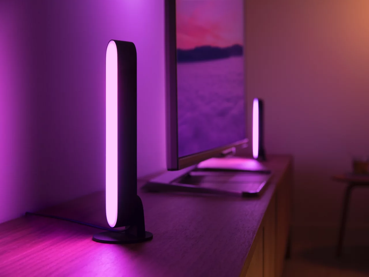Ein Raum mit einem Fernseher und zwei schwarzen Lampen mit rosa Licht auf einer Holzoberfläche. Die Wand ist lila.