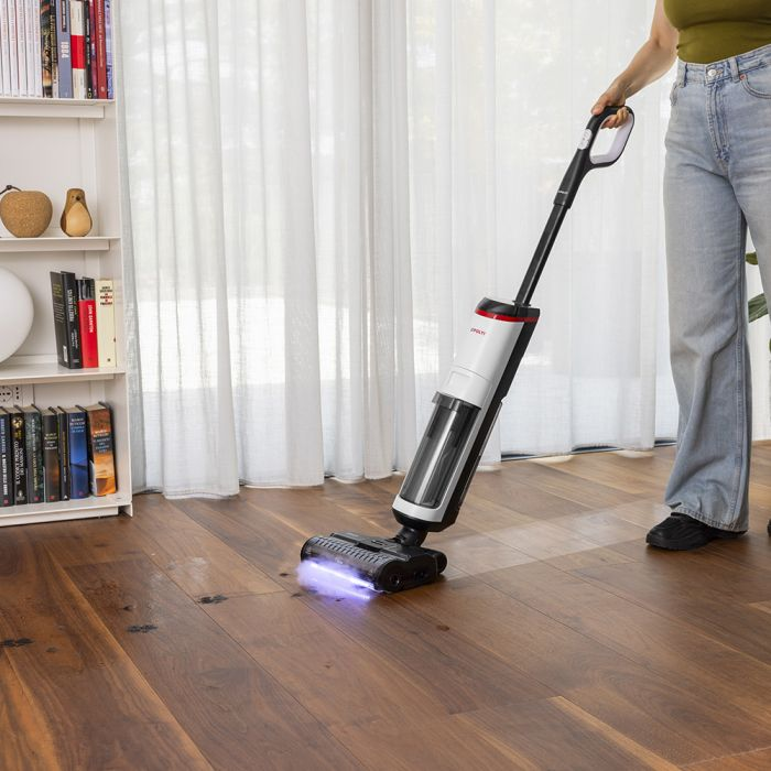 Una mujer usa una aspiradora en un suelo de madera. Una estantería está a la izquierda. Cortinas blancas en el fondo.