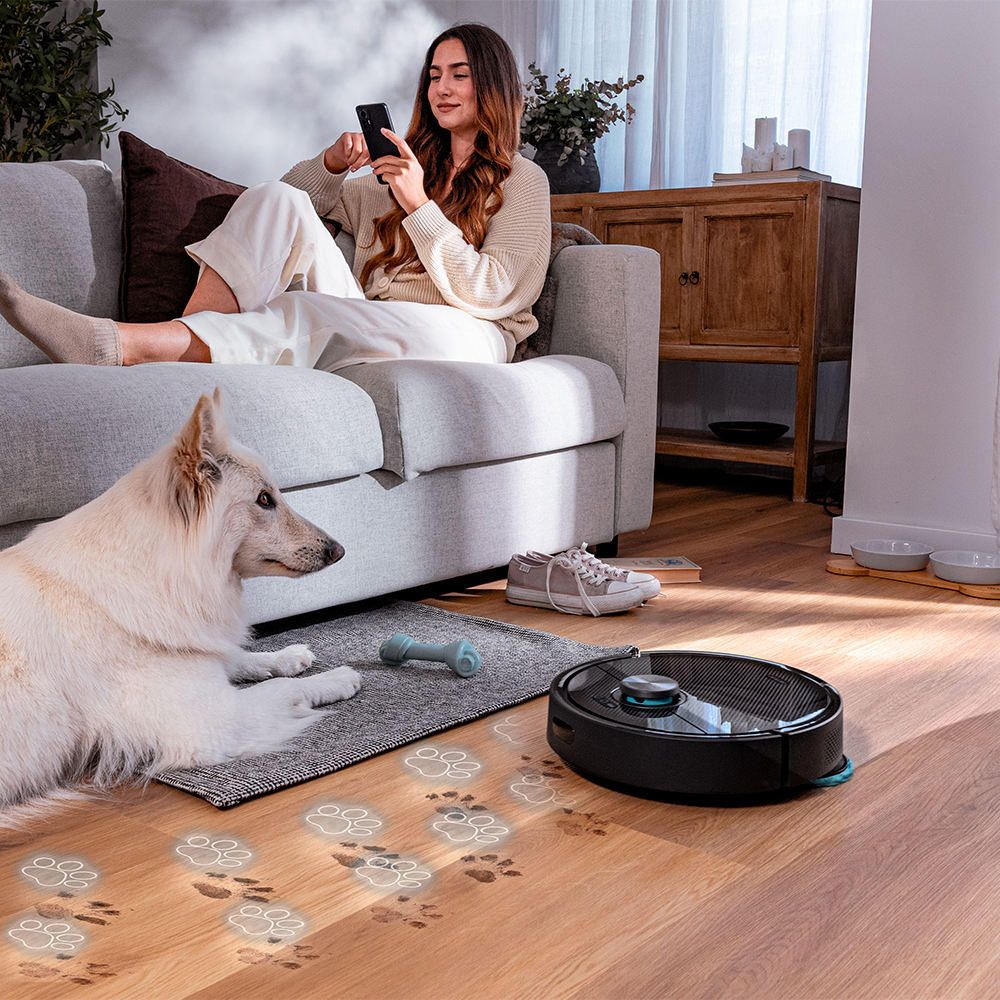 Mujer en sofá con teléfono, perro blanco, aspiradora robot, huellas de patas en el suelo. Zapatillas y hueso de juguete en el suelo.