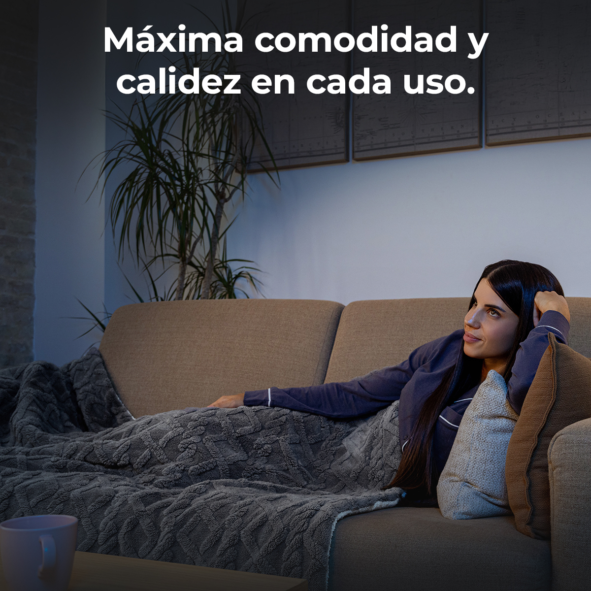 Una mujer se relaja en un sofá, cubierta por una manta. Está mirando hacia arriba y sonriendo. El fondo tiene una planta y arte.