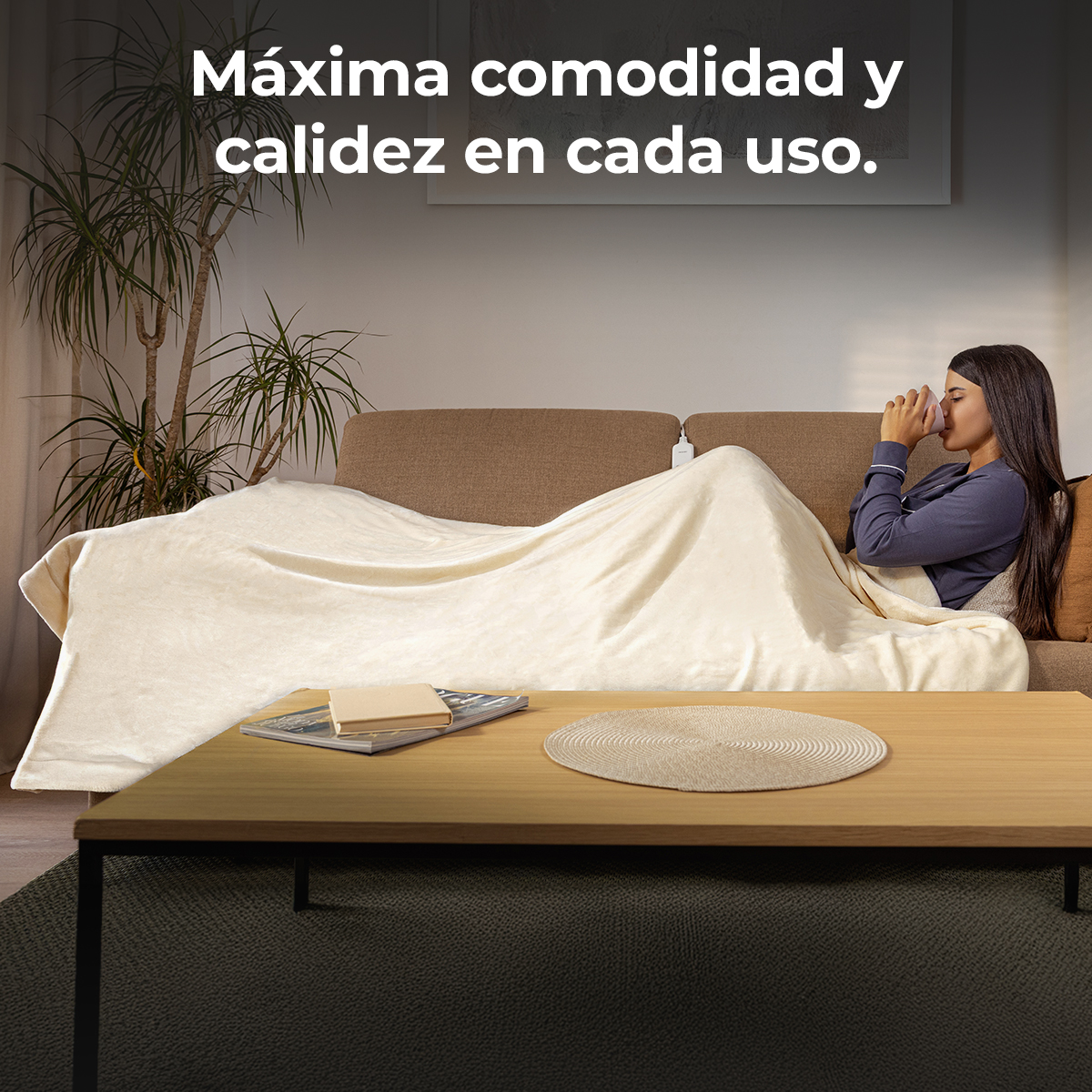Una mujer está sentada en un sofá cubierta con una manta. Bebe de una taza. Una mesa con un libro y un posavasos delante.