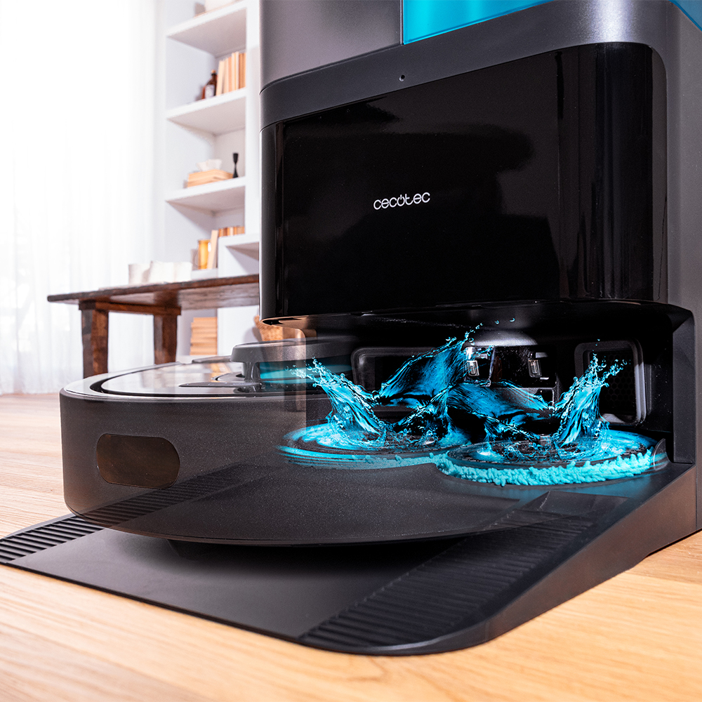Una aspiradora robot Cecotec negra con salpicaduras de agua, sobre suelo de madera. Fondo con mesa y estanterías.