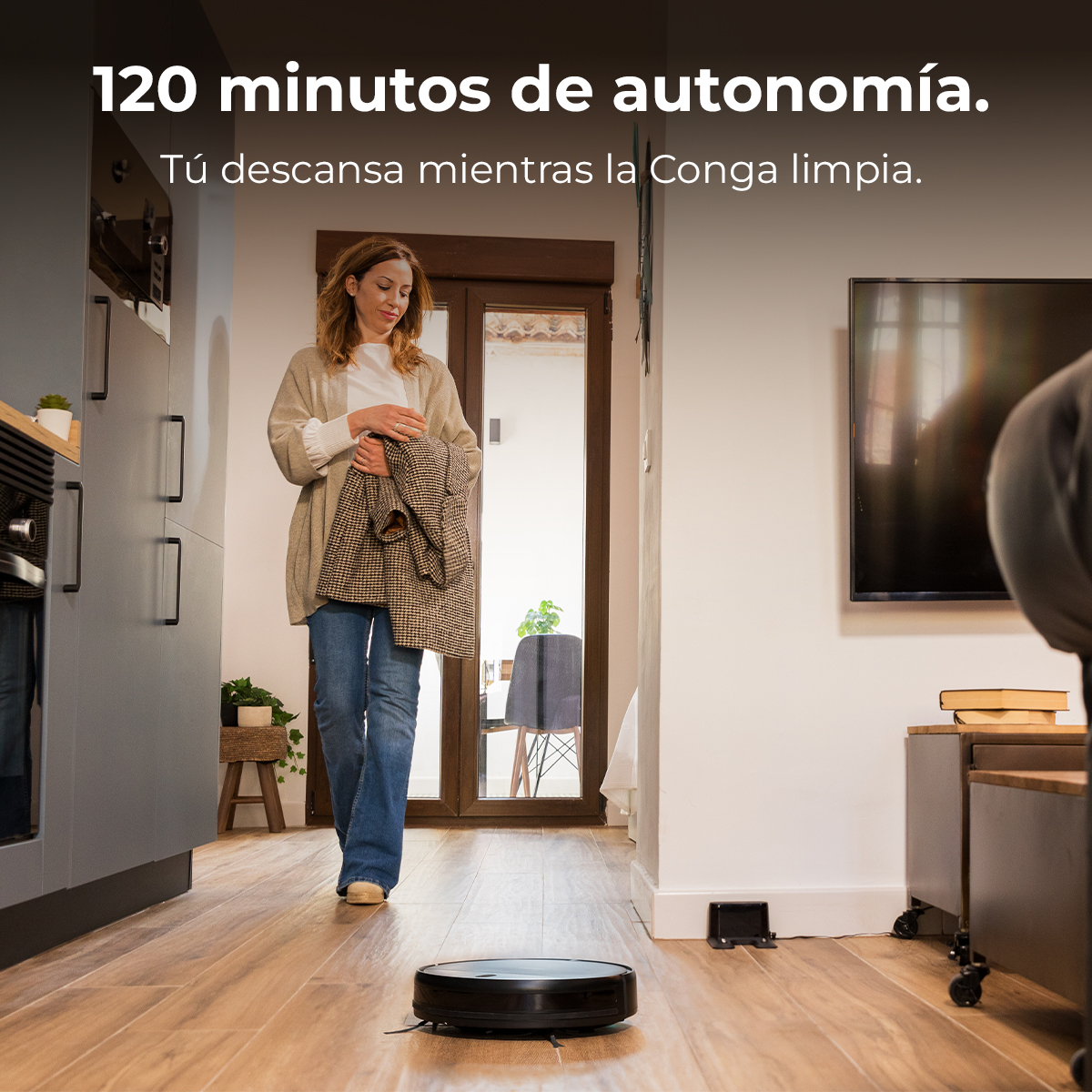 Mujer caminando con una chaqueta en casa. Un robot aspirador en el suelo. Texto: 120 minutos de autonomía.