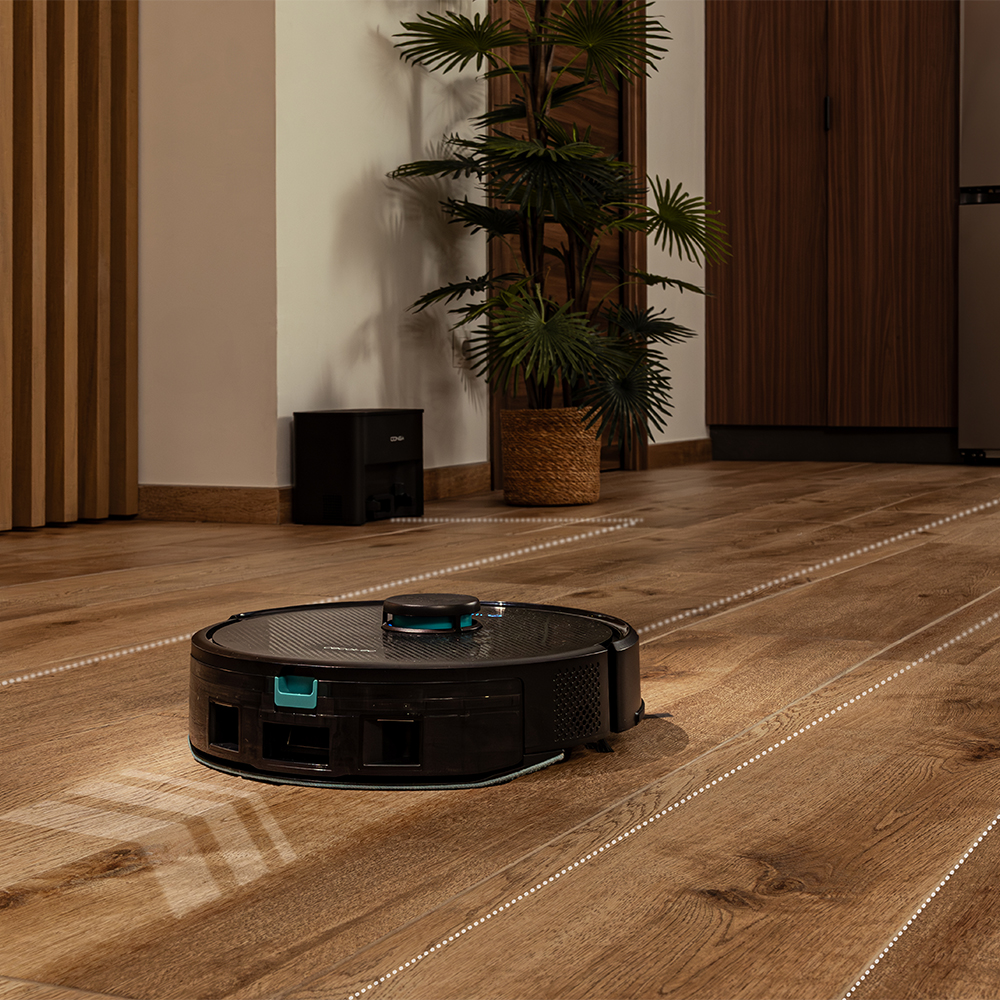 Una aspiradora robot en un suelo de madera. Una planta en una cesta y una estación de carga negra están en el fondo.
