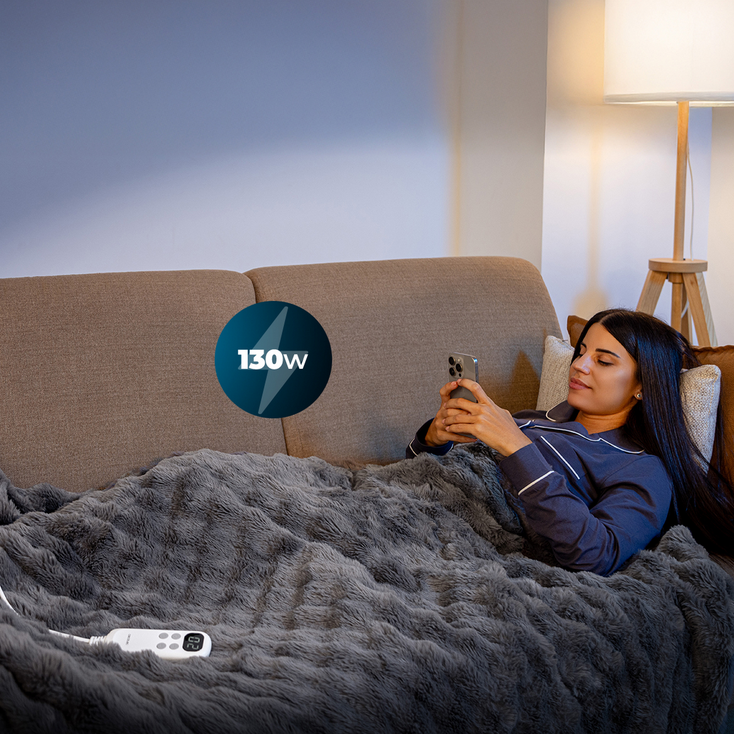 Mujer en pijama en el sofá, usando el teléfono, cubierta con una manta gris. Una lámpara de pie está detrás de ella. Icono de potencia de 130w presente.