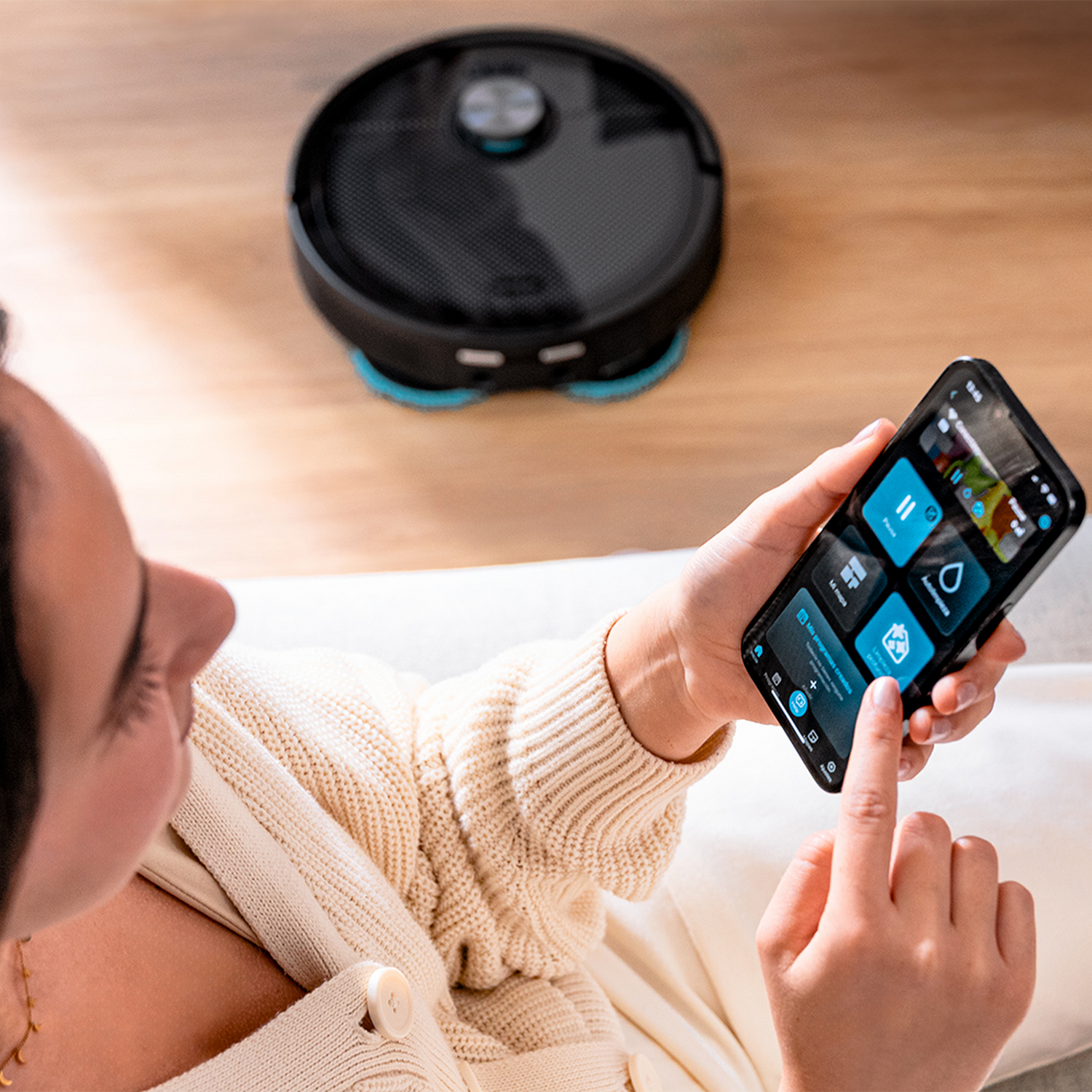 Una persona usa un smartphone para controlar una aspiradora robot. La pantalla del teléfono muestra controles de la aplicación. Una aspiradora negra está en el suelo.