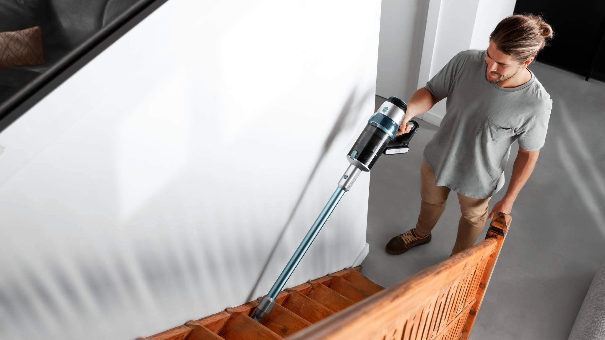 Un hombre aspira escaleras de madera con una aspiradora inalámbrica. Viste camisa gris y pantalones color caqui.