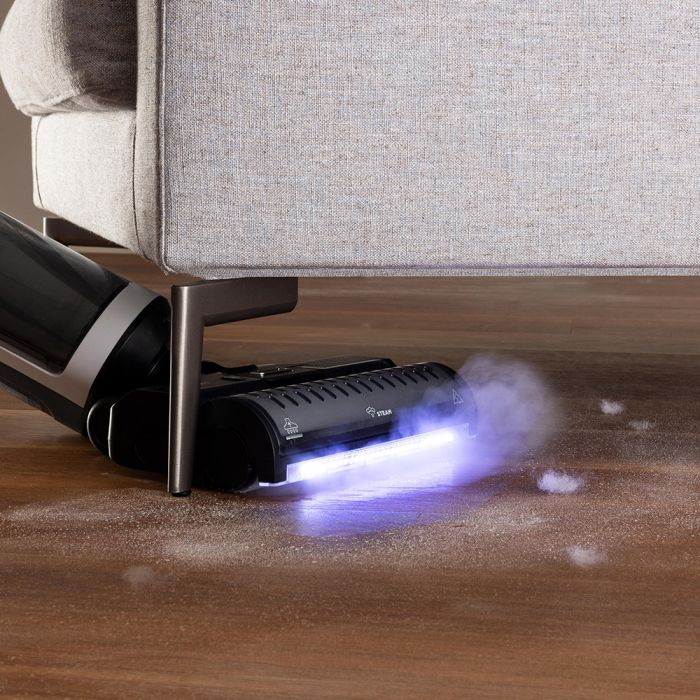 Una limpiadora de vapor negra está limpiando debajo de un sofá gris. Se ven partículas de vapor y polvo en el suelo de madera. La máquina tiene una luz azul.