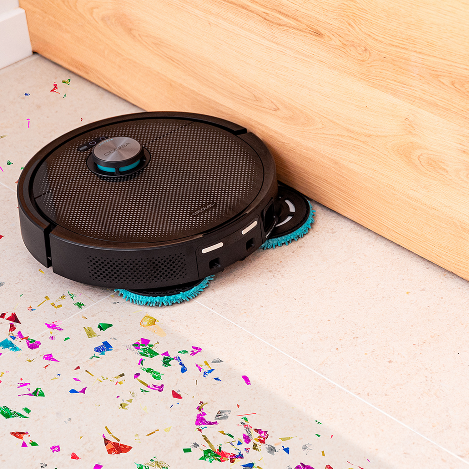 Una aspiradora robot negra está en un suelo embaldosado lleno de confeti, junto a una pared de madera.