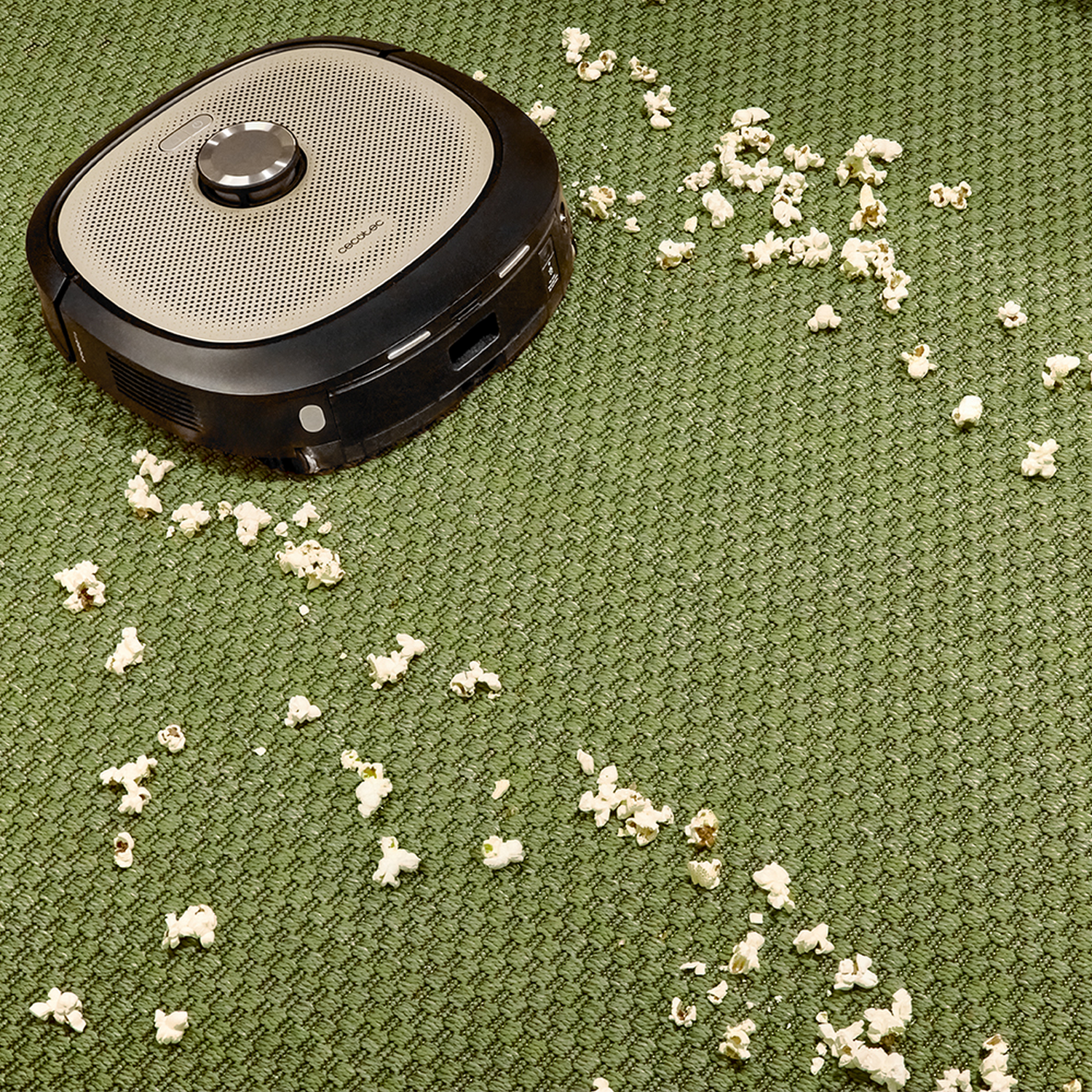 Una aspiradora robot en una alfombra verde, rodeada de palomitas de maíz esparcidas. La aspiradora es negra y beige.