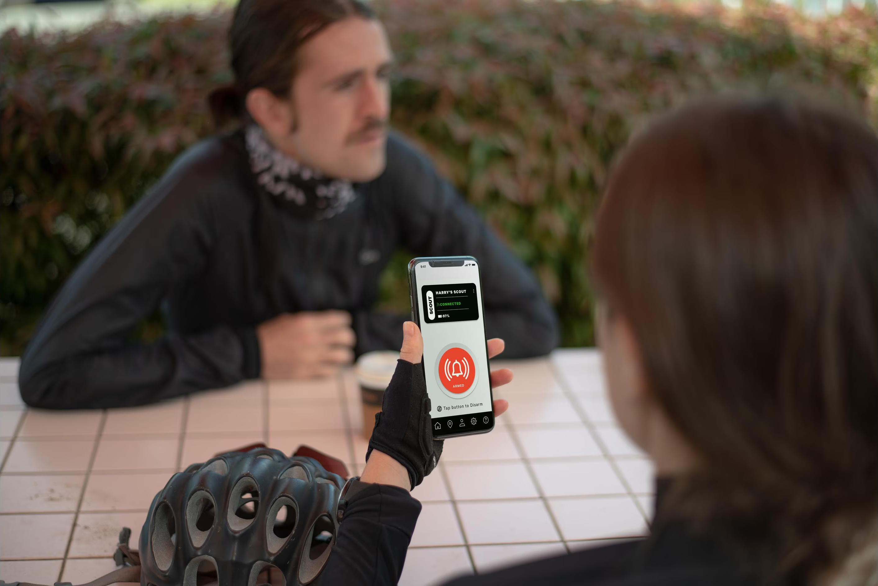 Eine Person hält ein Telefon mit der App 'Harry's Scout' und dem Status 'bewaffnet'. Eine andere Person ist im Hintergrund.