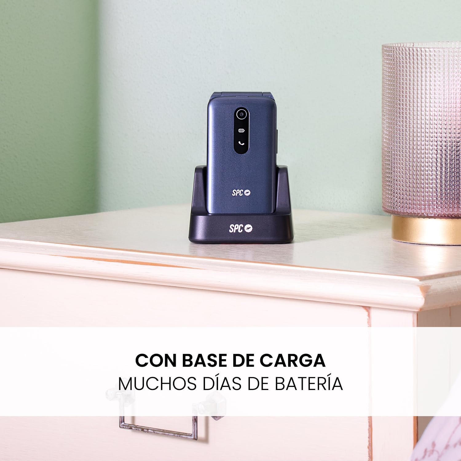 Un teléfono plegable azul oscuro en su base de carga sobre una mesita de noche blanca. Lámpara rosa texturizada a la derecha. Fondo verde.
