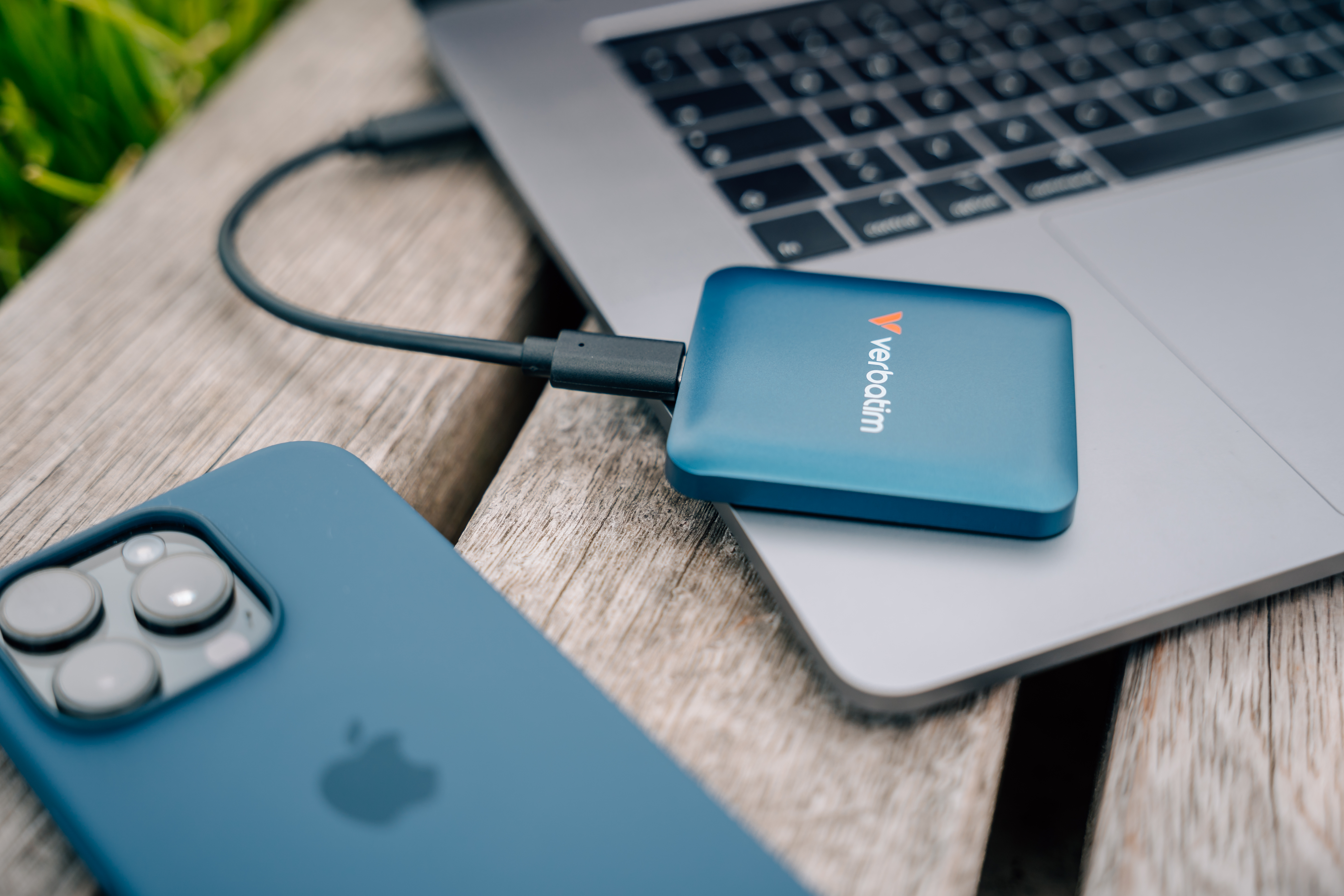 Blaue externe Verbatim-Festplatte, an Laptop angeschlossen. Ein iPhone liegt daneben auf Holz.