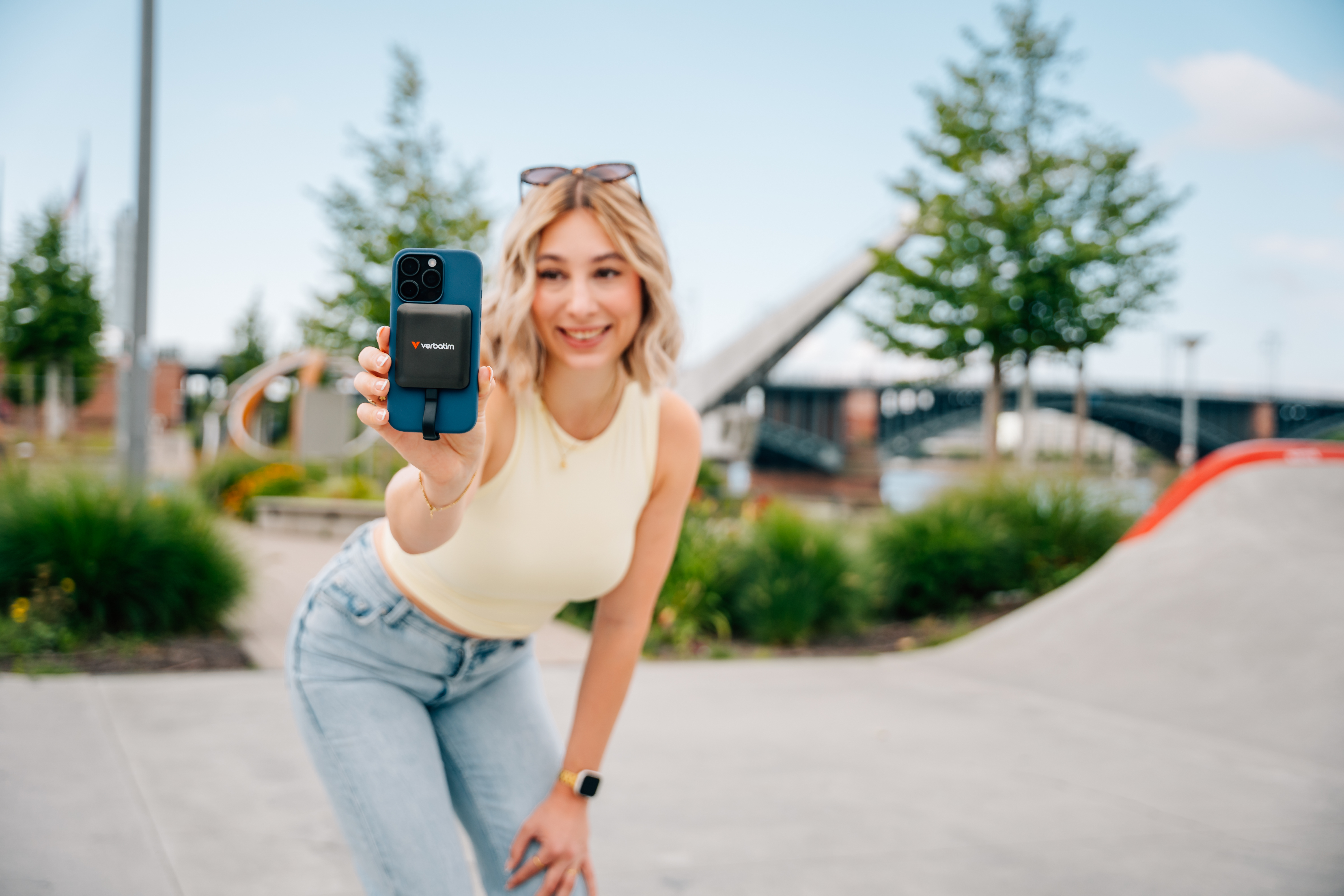 Eine Frau hält ein Telefon mit Ladegerät hoch. Im Hintergrund Bäume, eine Brücke und ein Skatepark.