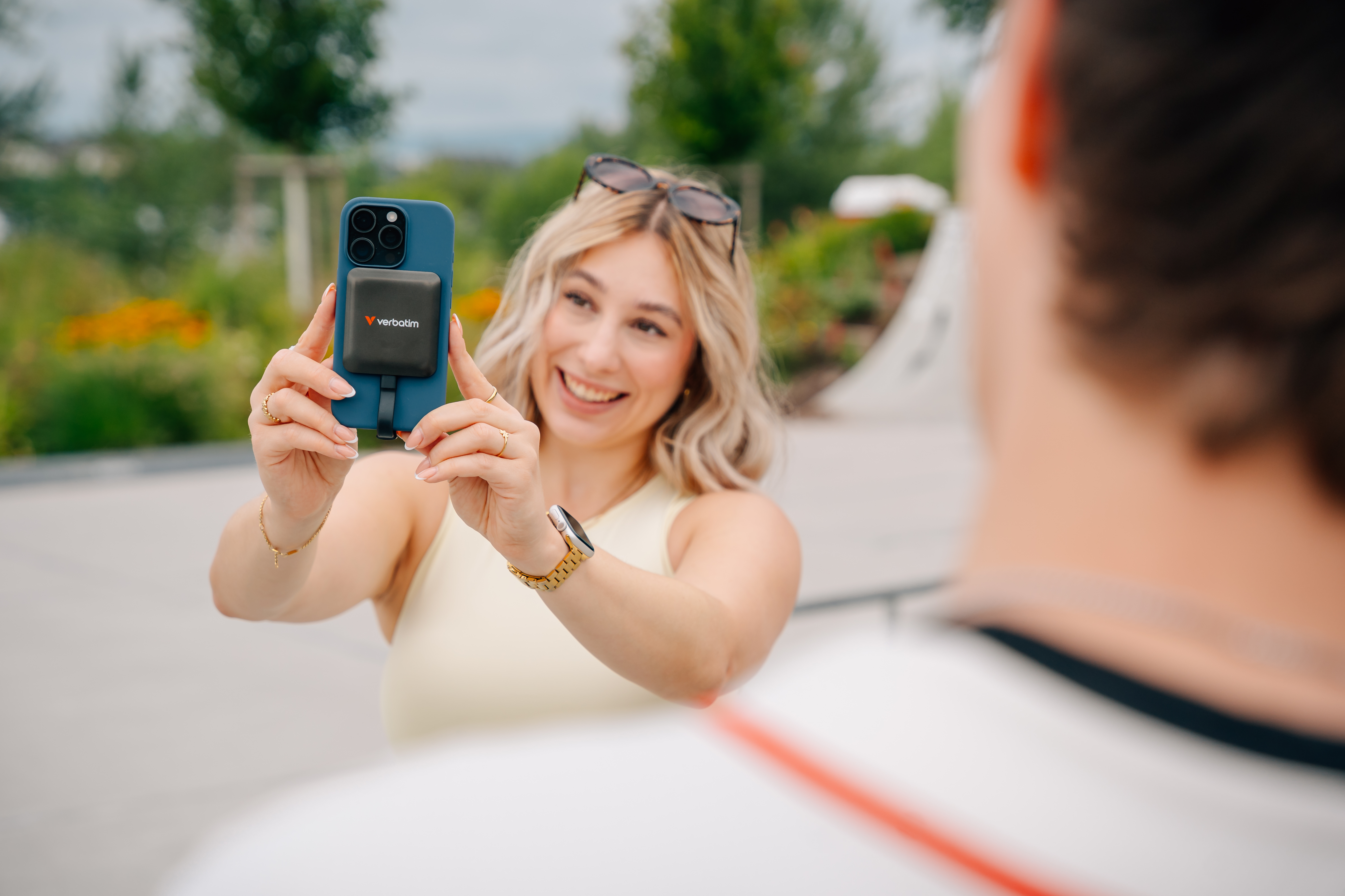 Frau macht Selfie mit Telefon, Powerbank befestigt. Sonnenbrille auf dem Kopf. Person im Vordergrund.