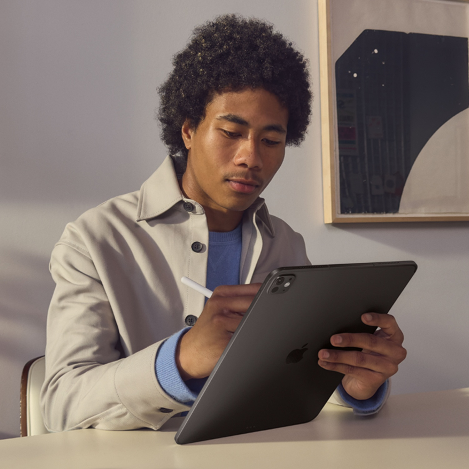 Un uomo con i capelli ricci usa un tablet e una penna. Indossa una giacca leggera e un maglione blu. Un quadro è appeso al muro.