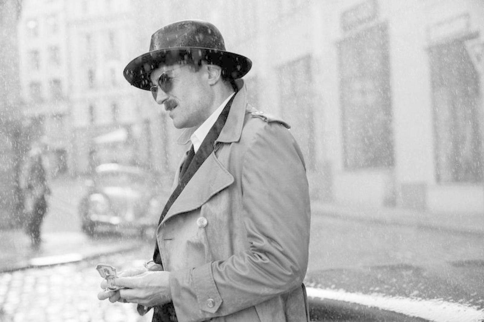 Mann mit Hut und Trenchcoat im Regen, hält einen kleinen Gegenstand. Im Hintergrund Gebäude, ein Auto und eine verschwommene Figur.