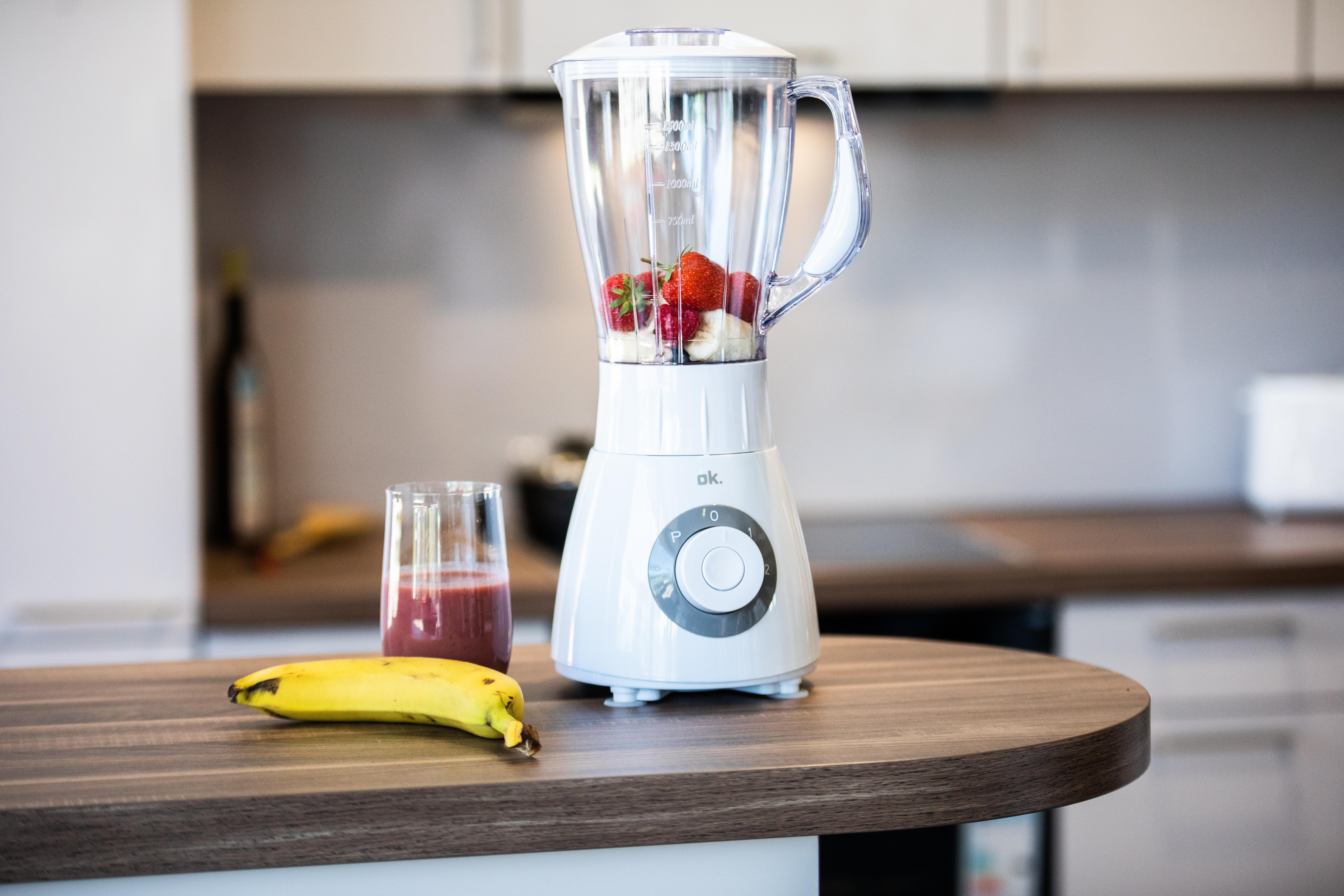 Biały blender z owocami w środku, szklanka smoothie i banan na drewnianej powierzchni.