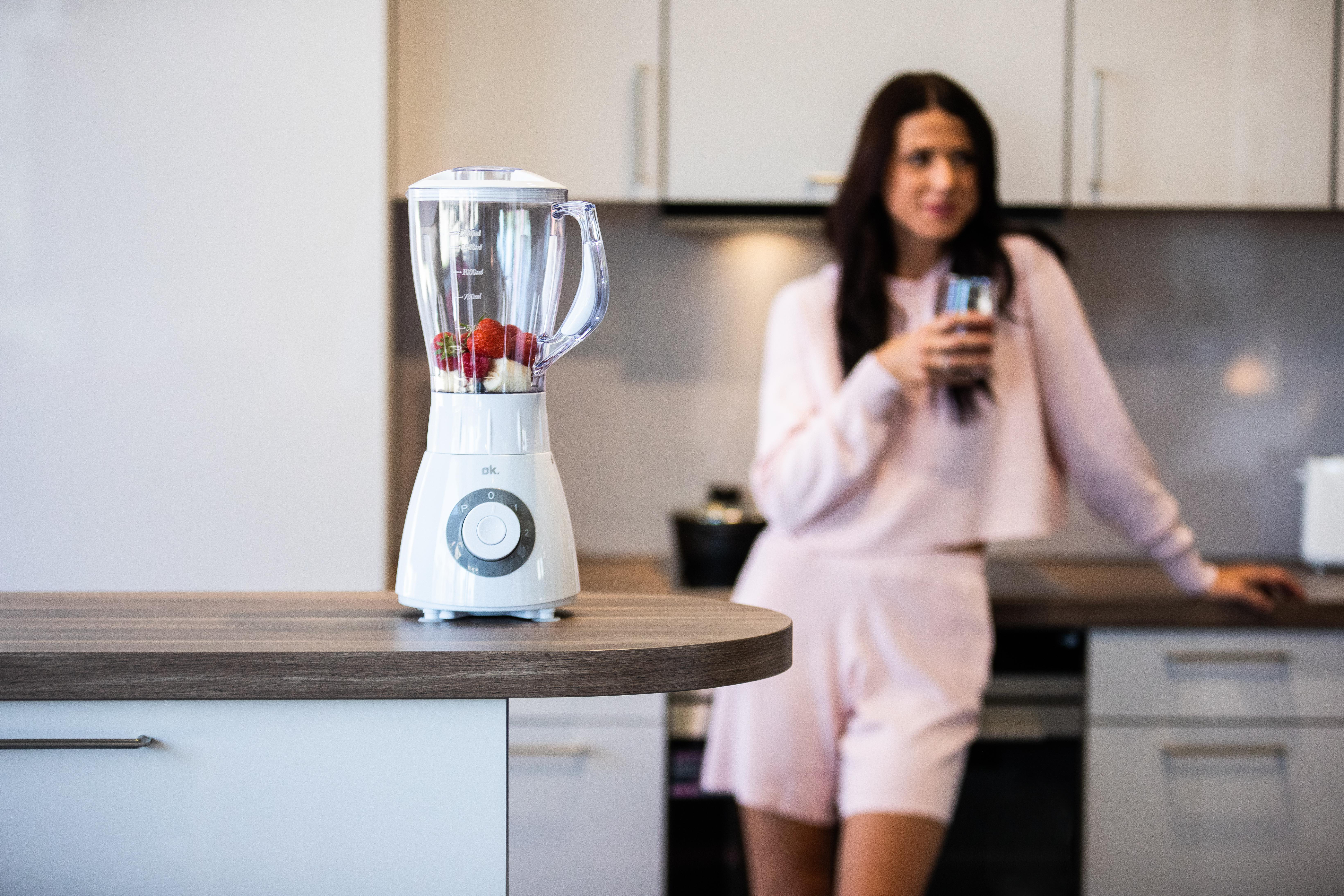 Biały blender z truskawkami i kobieta w różowej piżamie trzymająca szklankę, w kuchni.