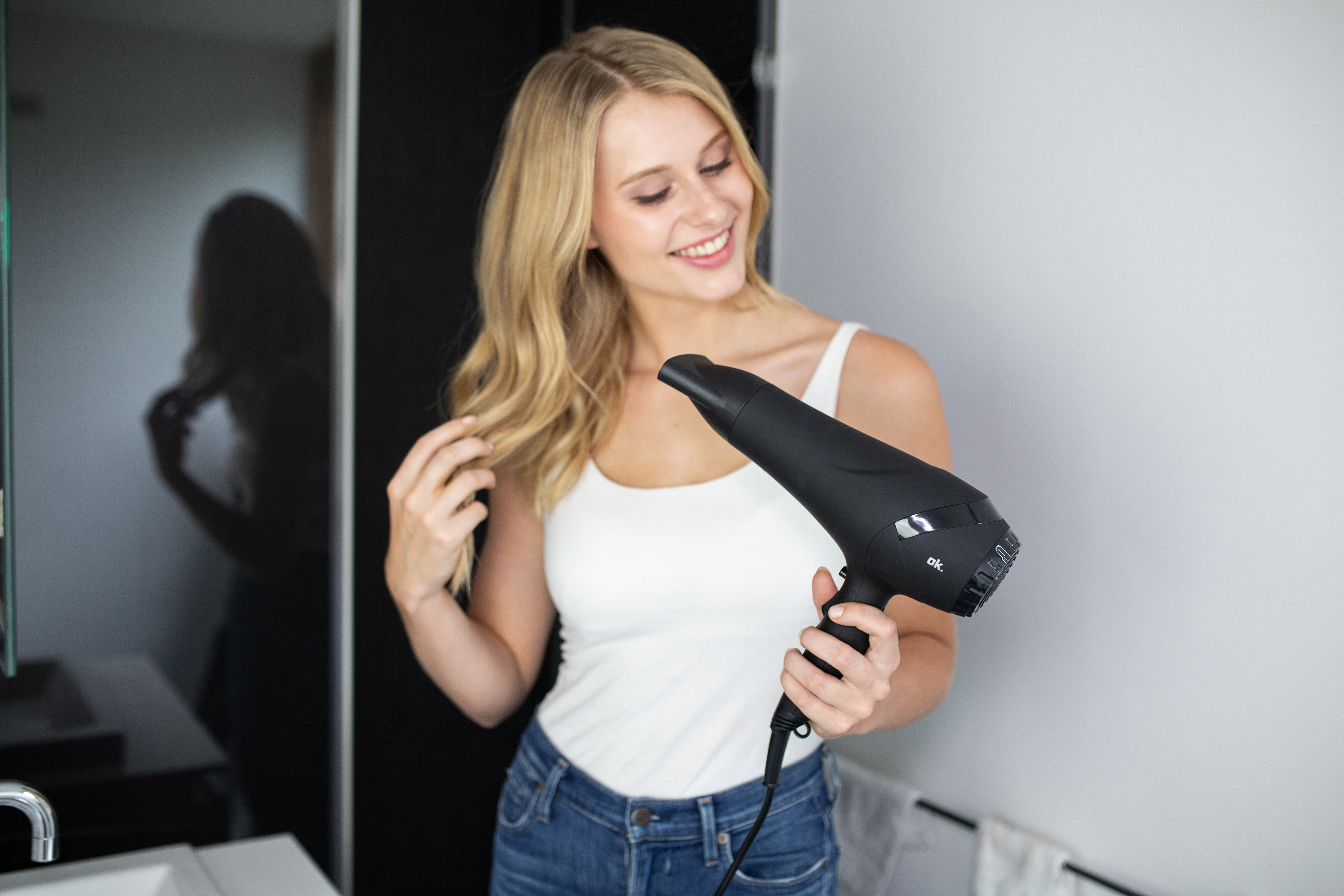 Kobieta z blond włosami trzyma czarną suszarkę do włosów. Uśmiecha się, nosząc biały top i dżinsy w łazience.