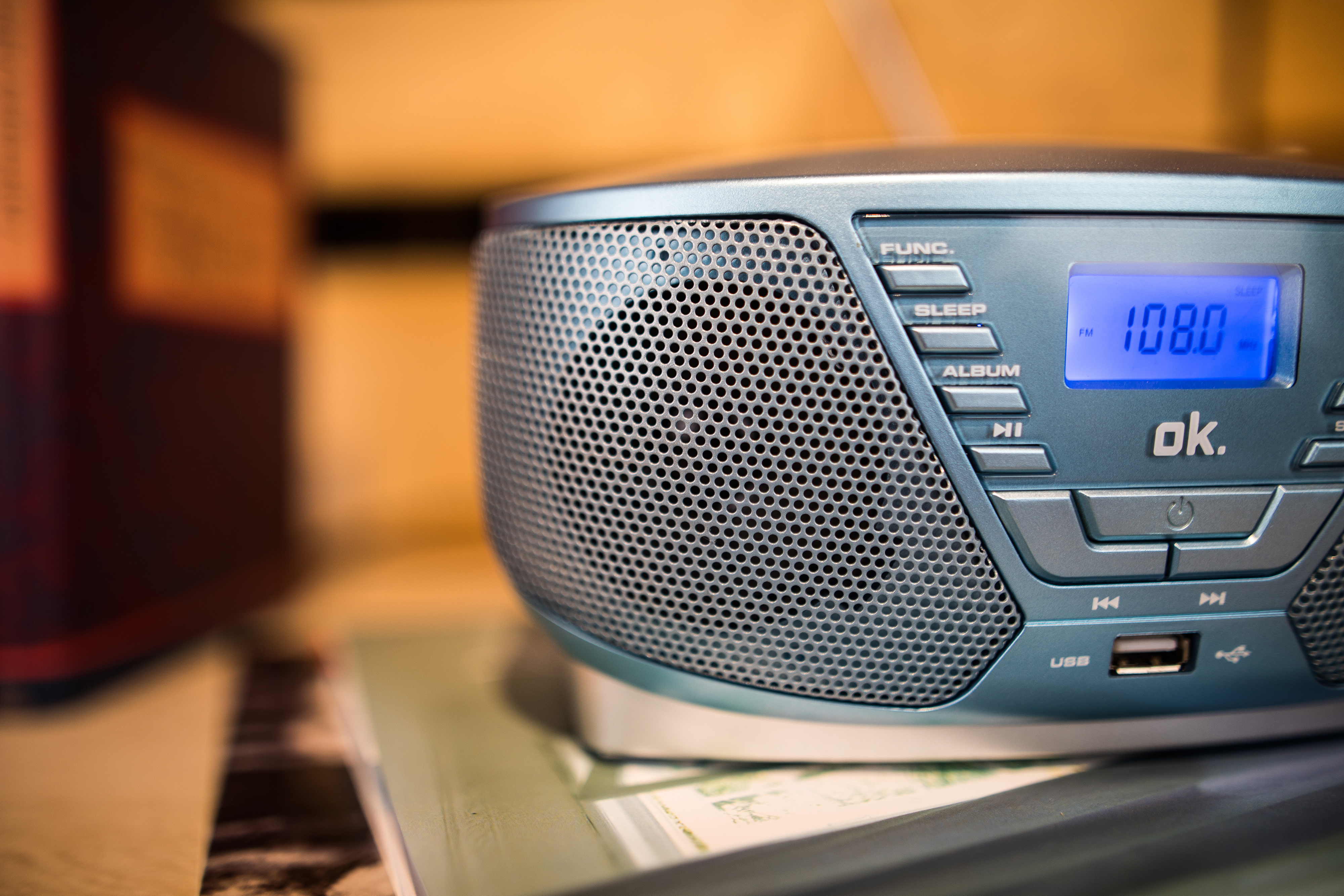 Ein blaues Radio mit Anzeige von 108,0 liegt auf einem Buch. Ein braunes Objekt ist im Hintergrund unscharf.