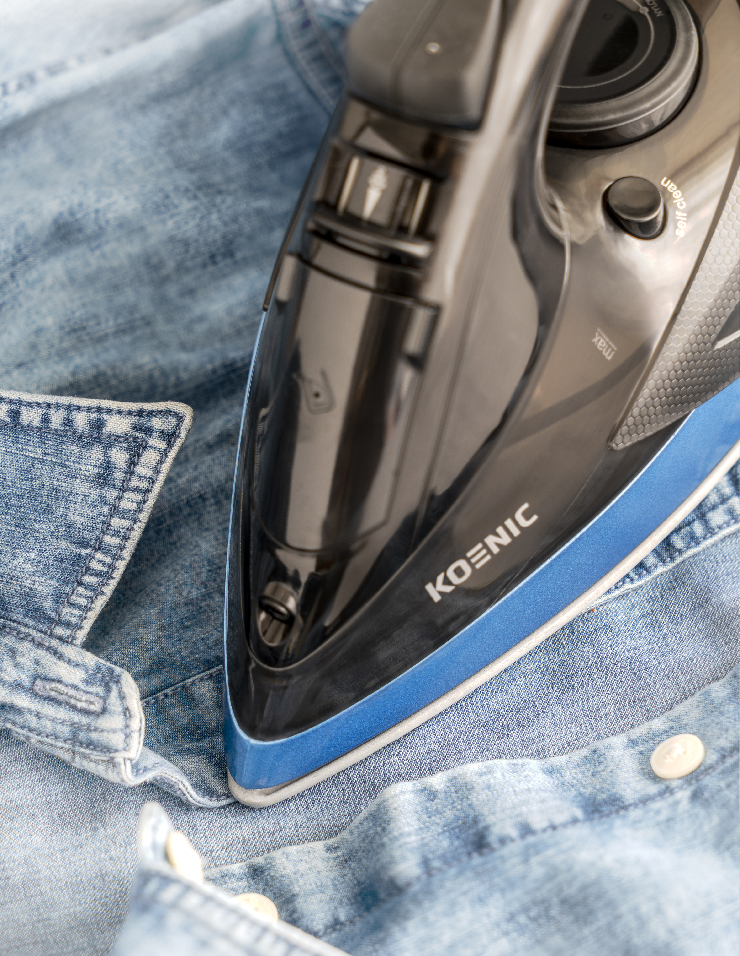 Ein schwarzes Bügeleisen liegt auf einem blauen Jeanshemd. Das Bügeleisen hat eine blaue Kante. KOENIC steht auf dem Bügeleisen.
