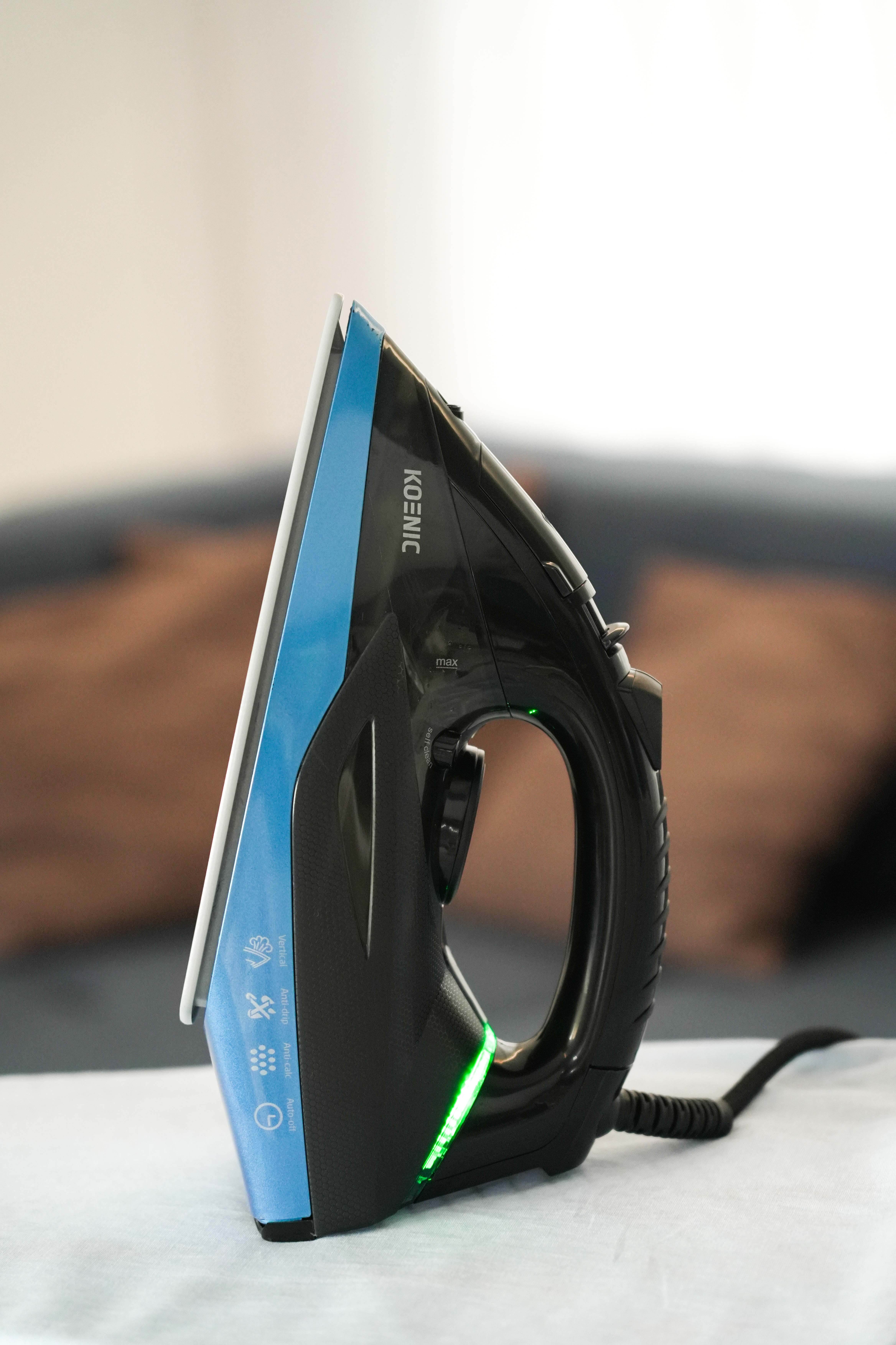 Ein schwarz-blaues Bügeleisen liegt auf einem weißen Tuch. Das Bügeleisen trägt den Markennamen 'KOENIC'. Ein grünes Licht leuchtet.