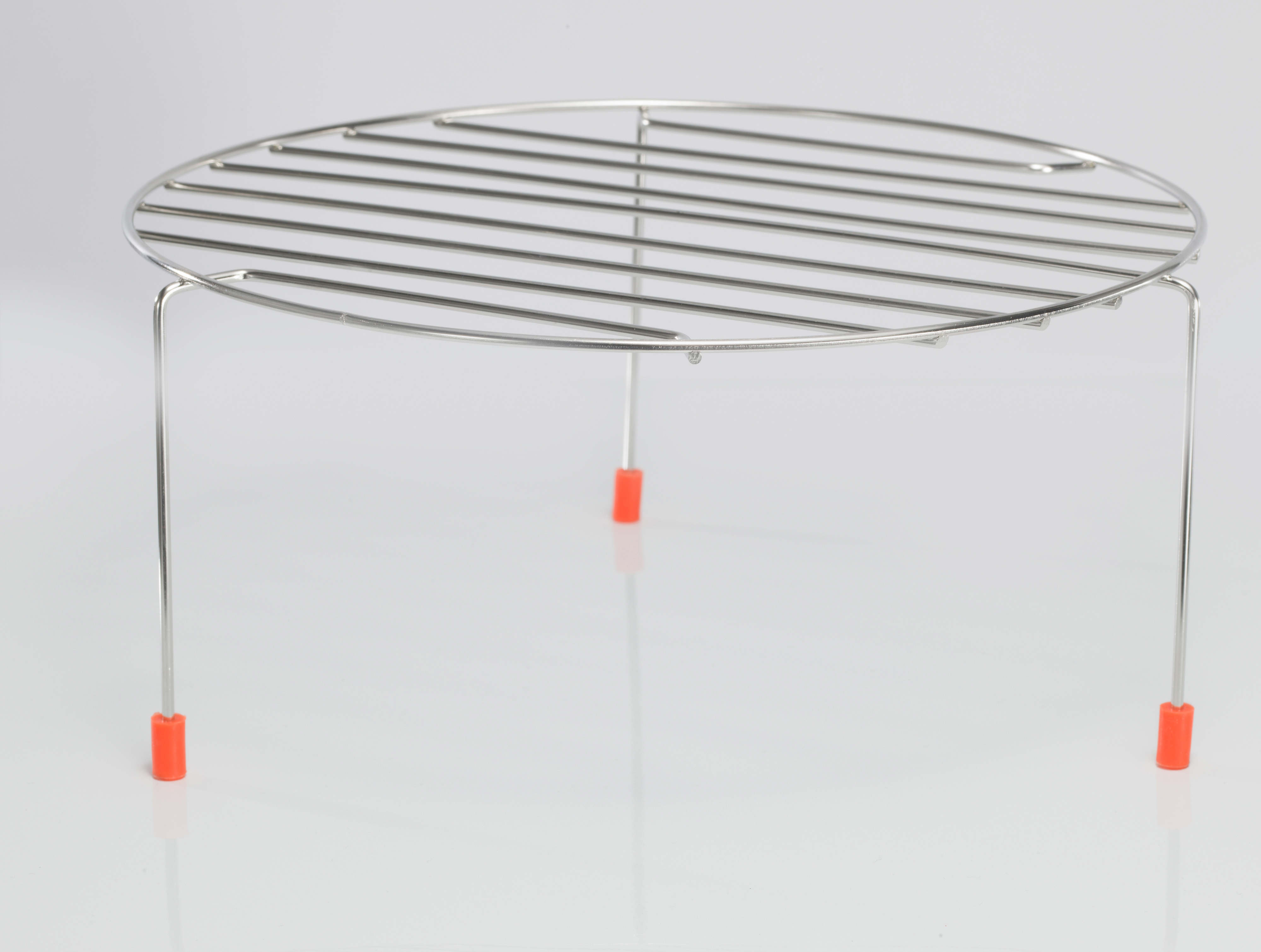 Okrągły, metalowy stojak do chłodzenia z trzema nóżkami. Nogi mają pomarańczowe gumowe końcówki. Na białej powierzchni.