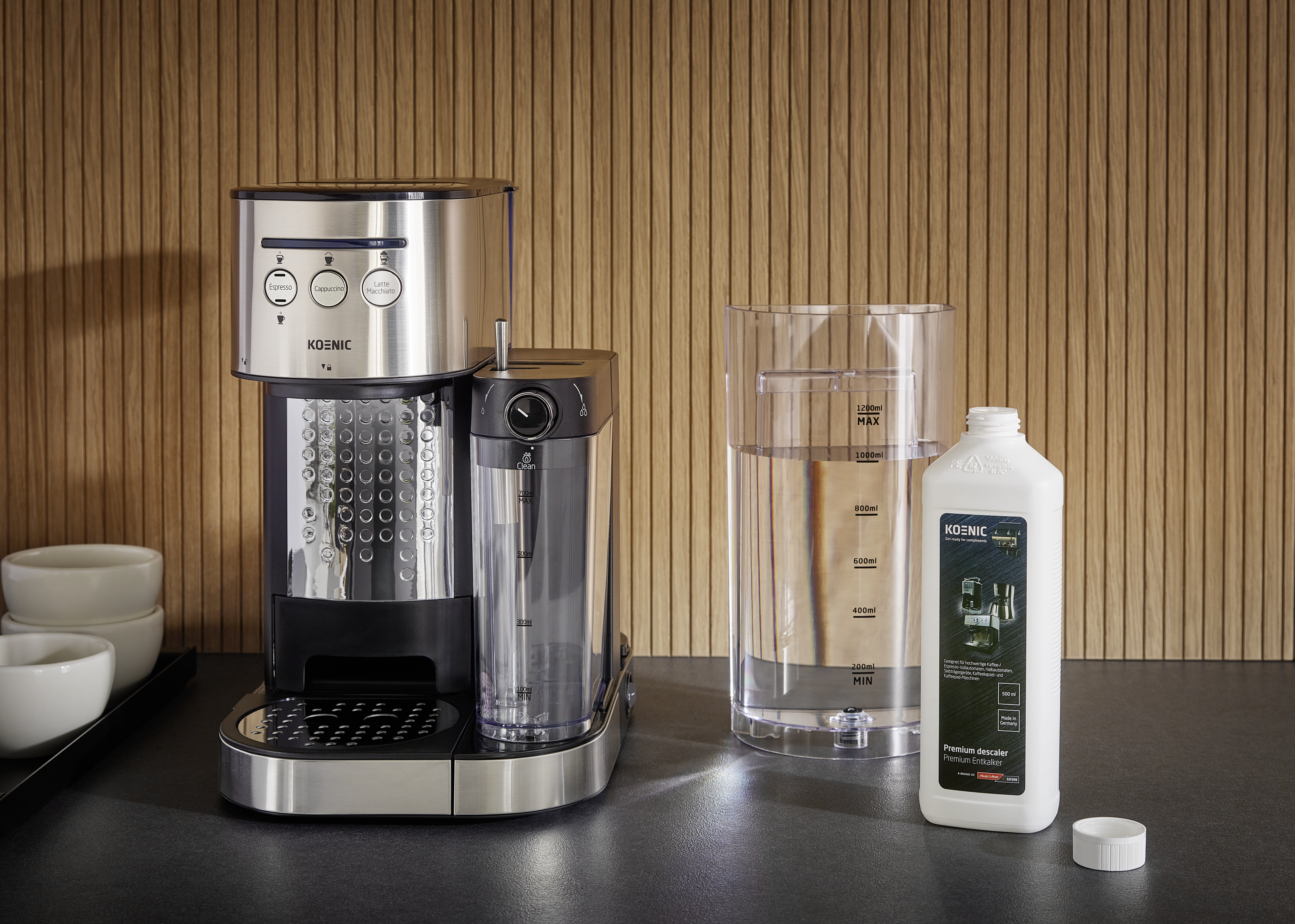 Kaffeemaschine, Wasserbehälter und Entkalkerflasche auf schwarzer Oberfläche vor Holzhintergrund.