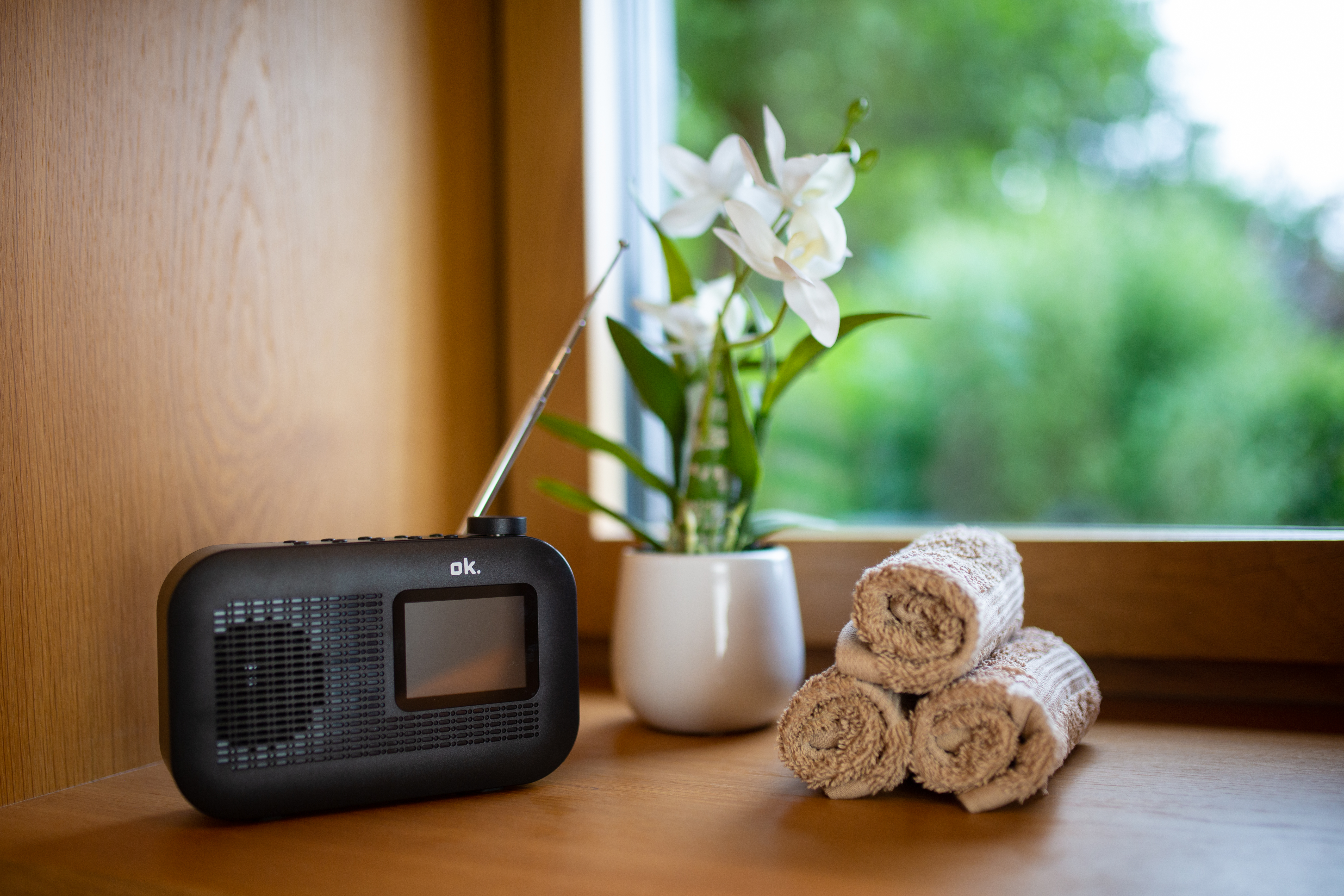 Ein schwarzes Radio, weiße Blumen und gerollte Handtücher liegen auf einer Holzoberfläche in der Nähe eines Fensters.