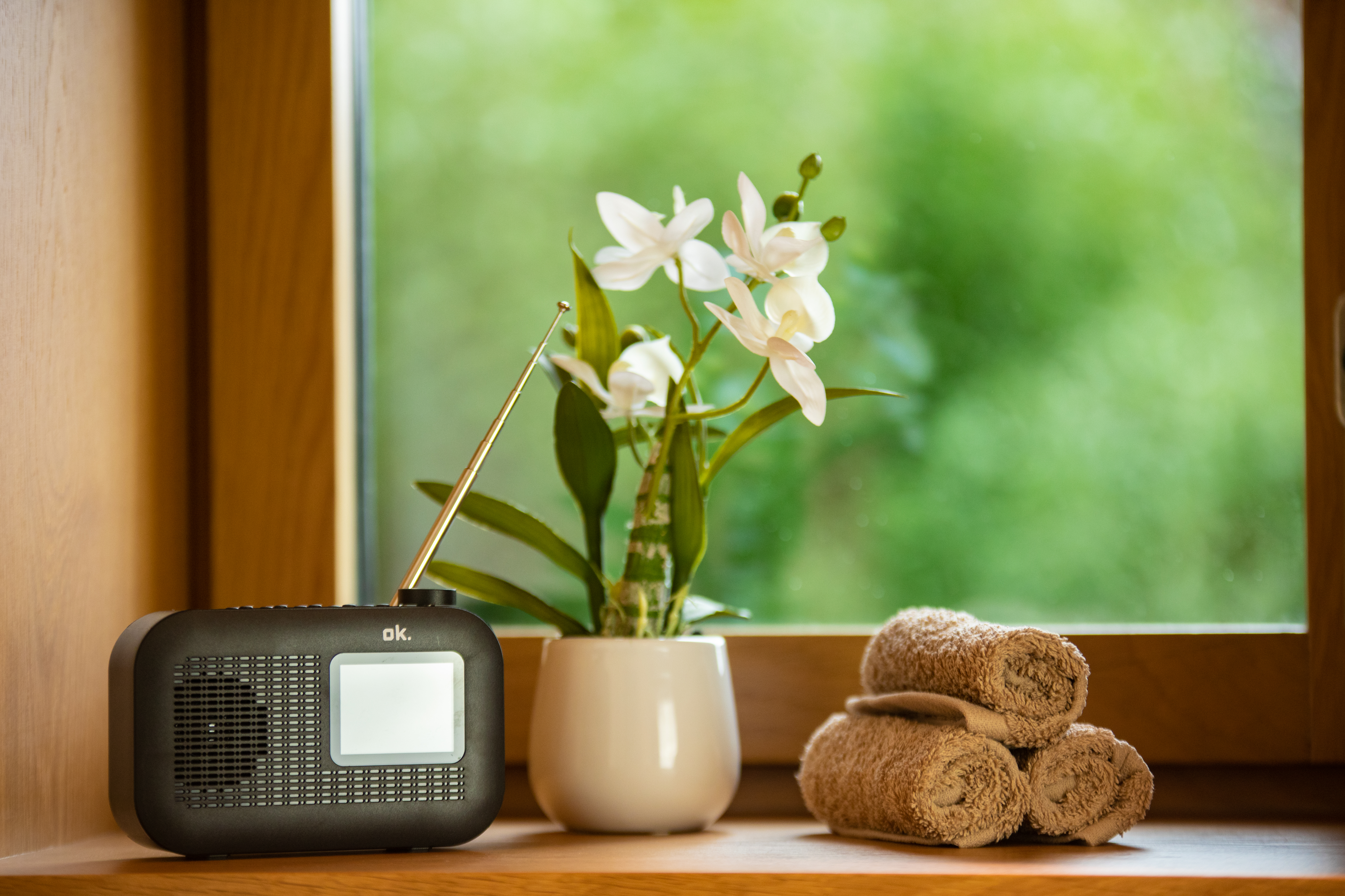 Ein schwarzes Radio, weiße Blumen in einer Vase und gerollte Handtücher stehen auf einer Holzoberfläche am Fenster.