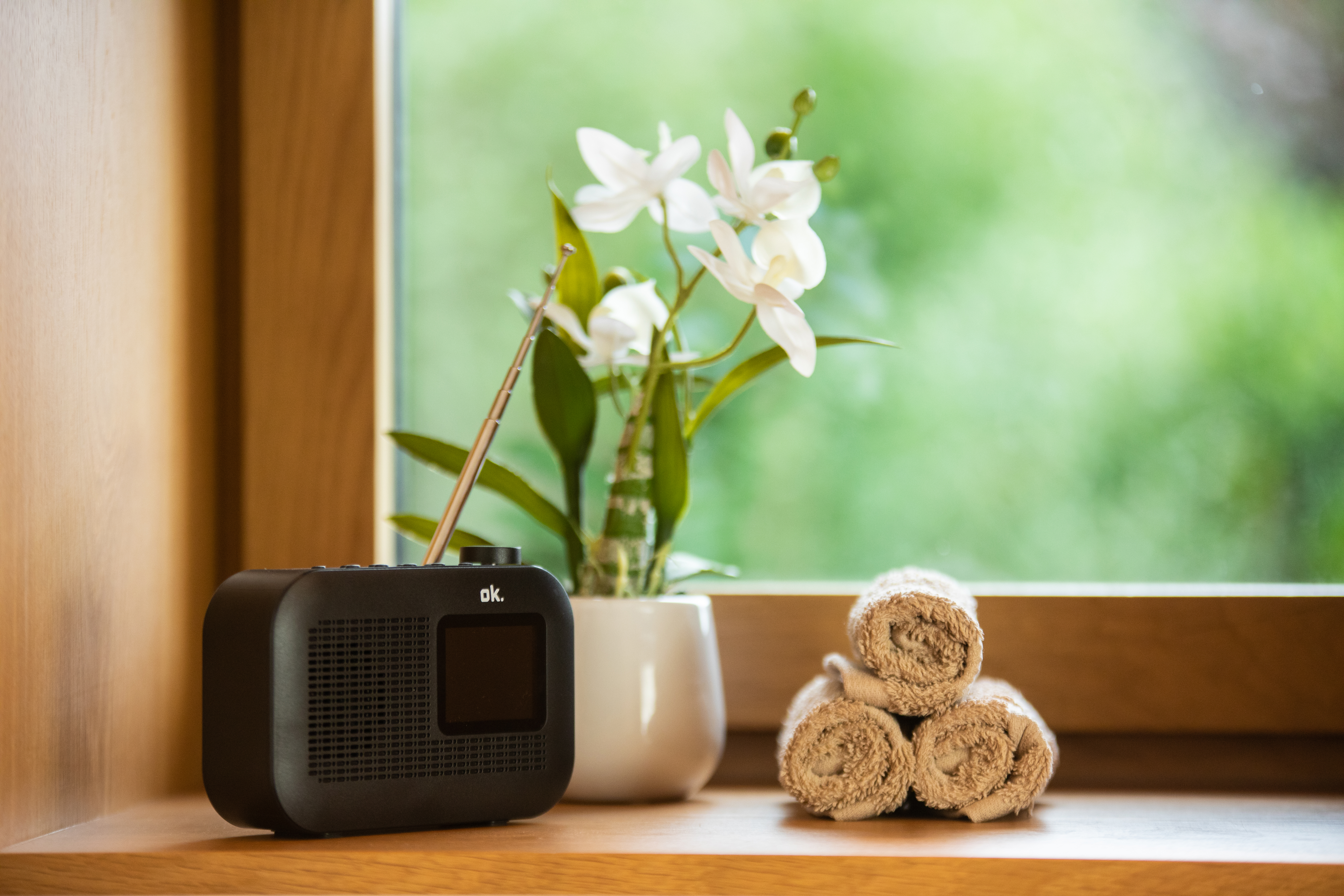 Ein schwarzes Radio, weiße Orchideen in einem Topf und gerollte Handtücher auf einer Holzoberfläche, mit grünem, verschwommenem Hintergrund.
