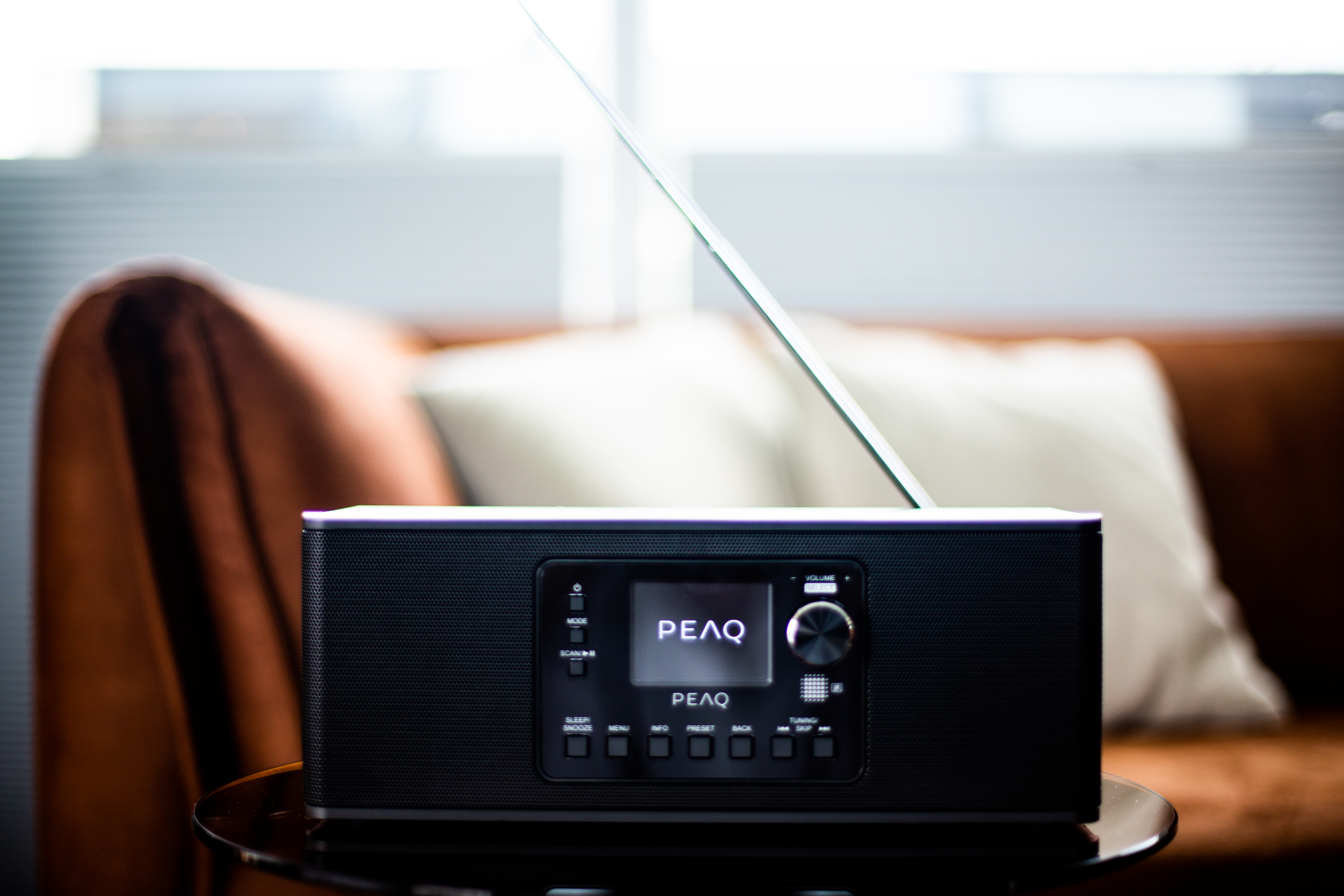 Czarny radio z anteną stoi na szklanym stole. W tle brązowa sofa.