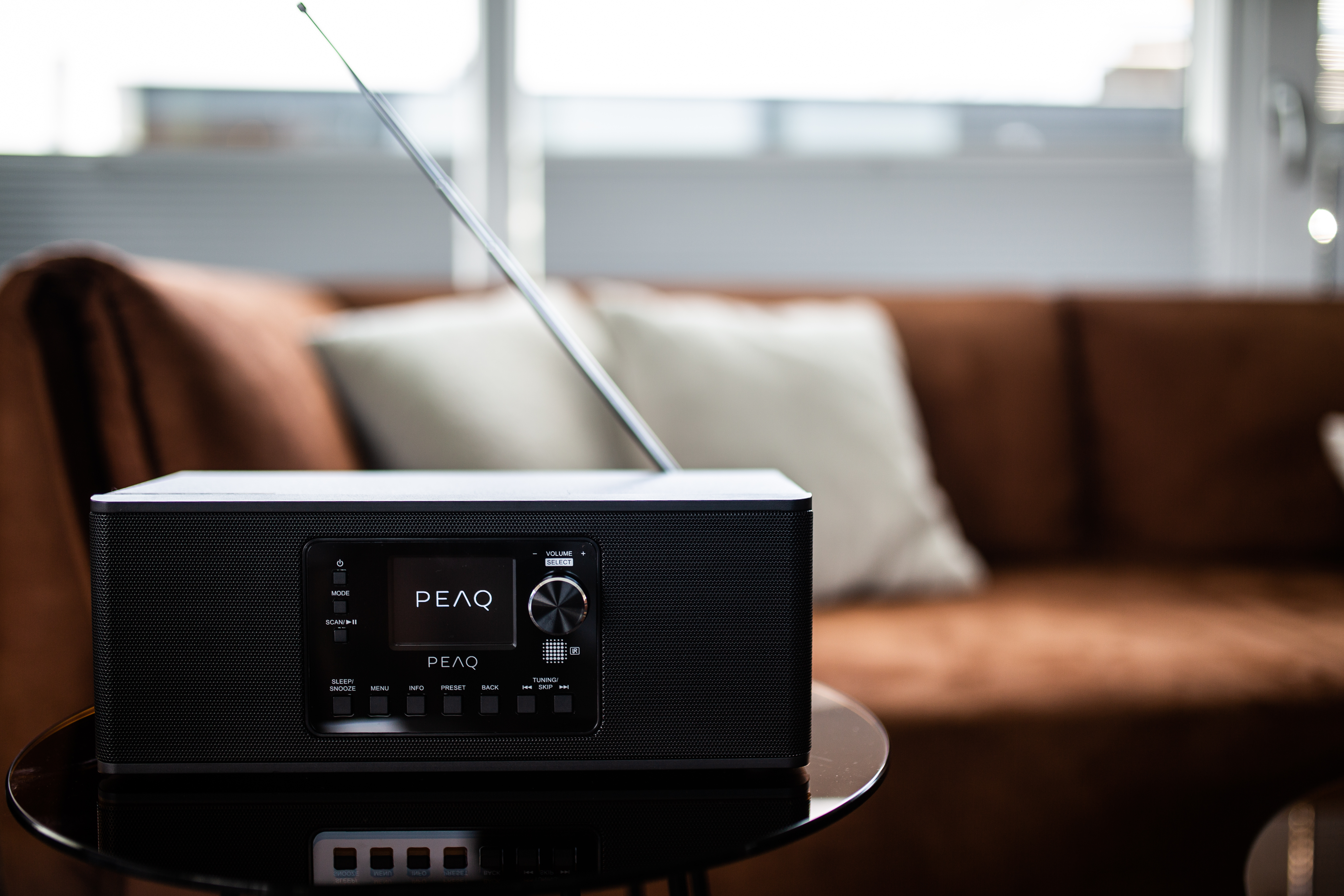 Czarny radioodbiornik z anteną stoi na szklanym stole. Brązowa sofa i biała poduszka w tle.