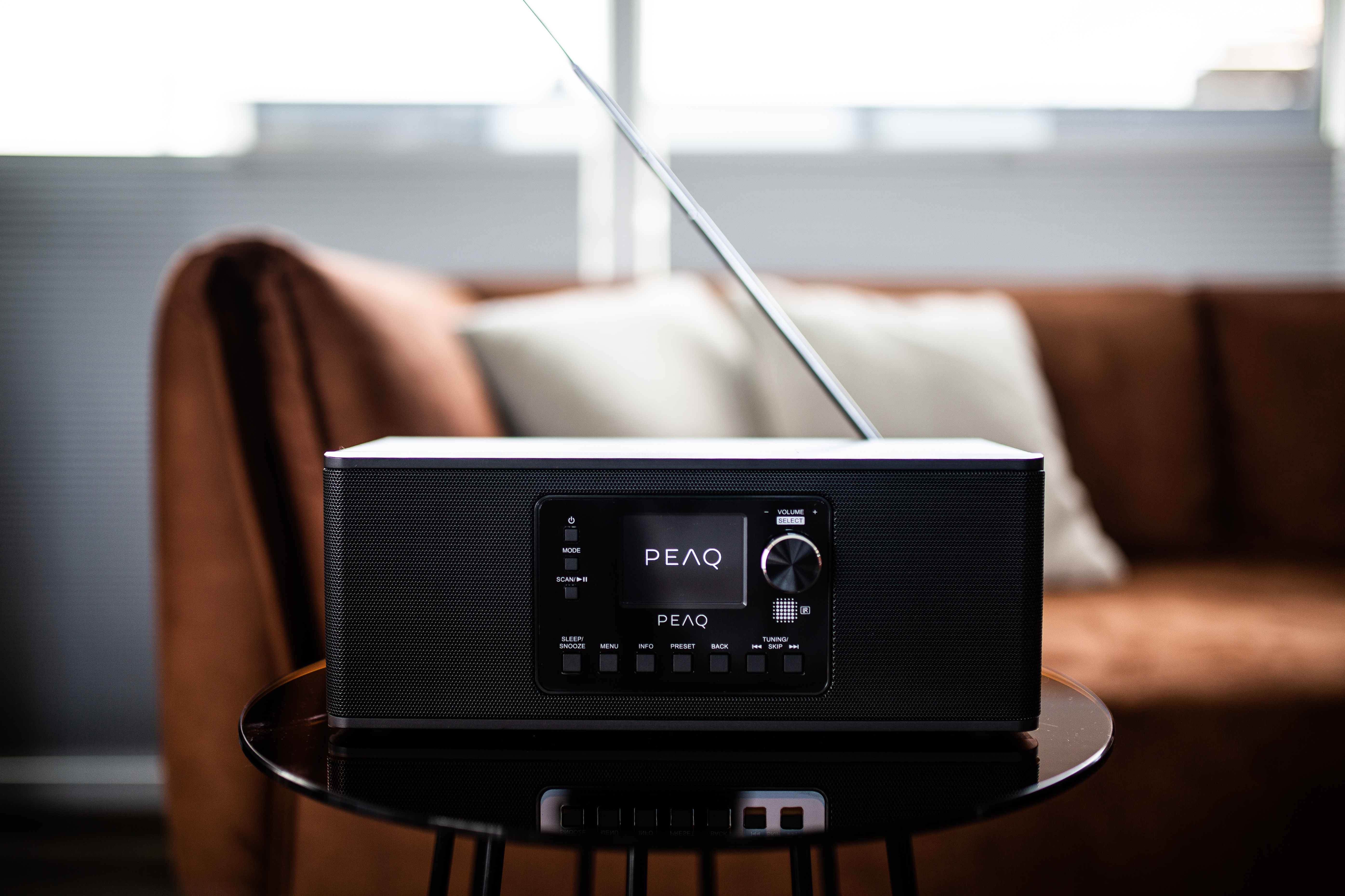 Czarny radioodtwarzacz PEAQ na czarnym stoliku z brązową sofą w tle. Antena wysunięta. Wyświetlacz pokazuje PEAQ.