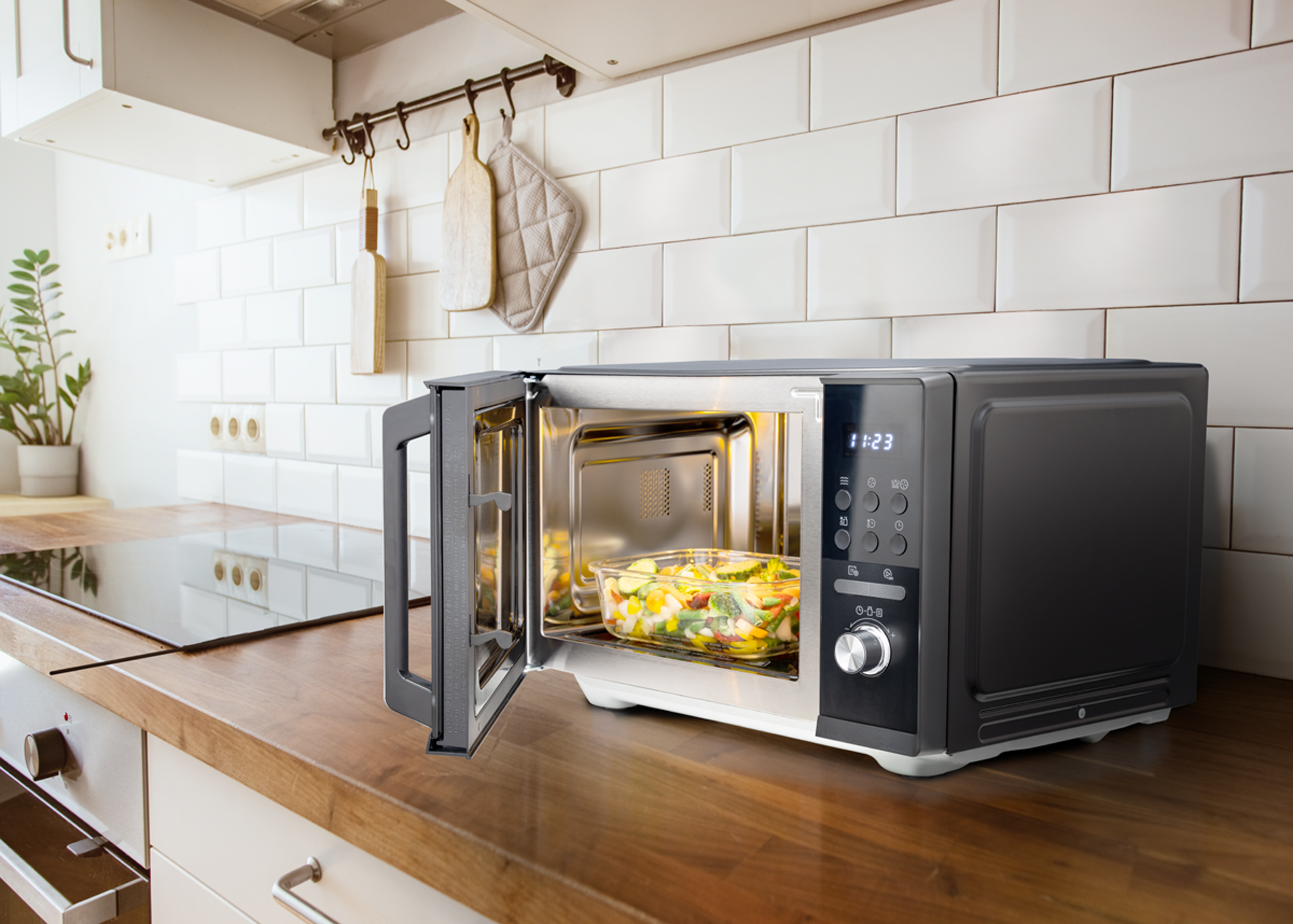 Mikrofalówka z otwartymi drzwiami, szklane naczynie z warzywami w środku. Kuchnia z białymi kafelkami i drewnianym blatem.