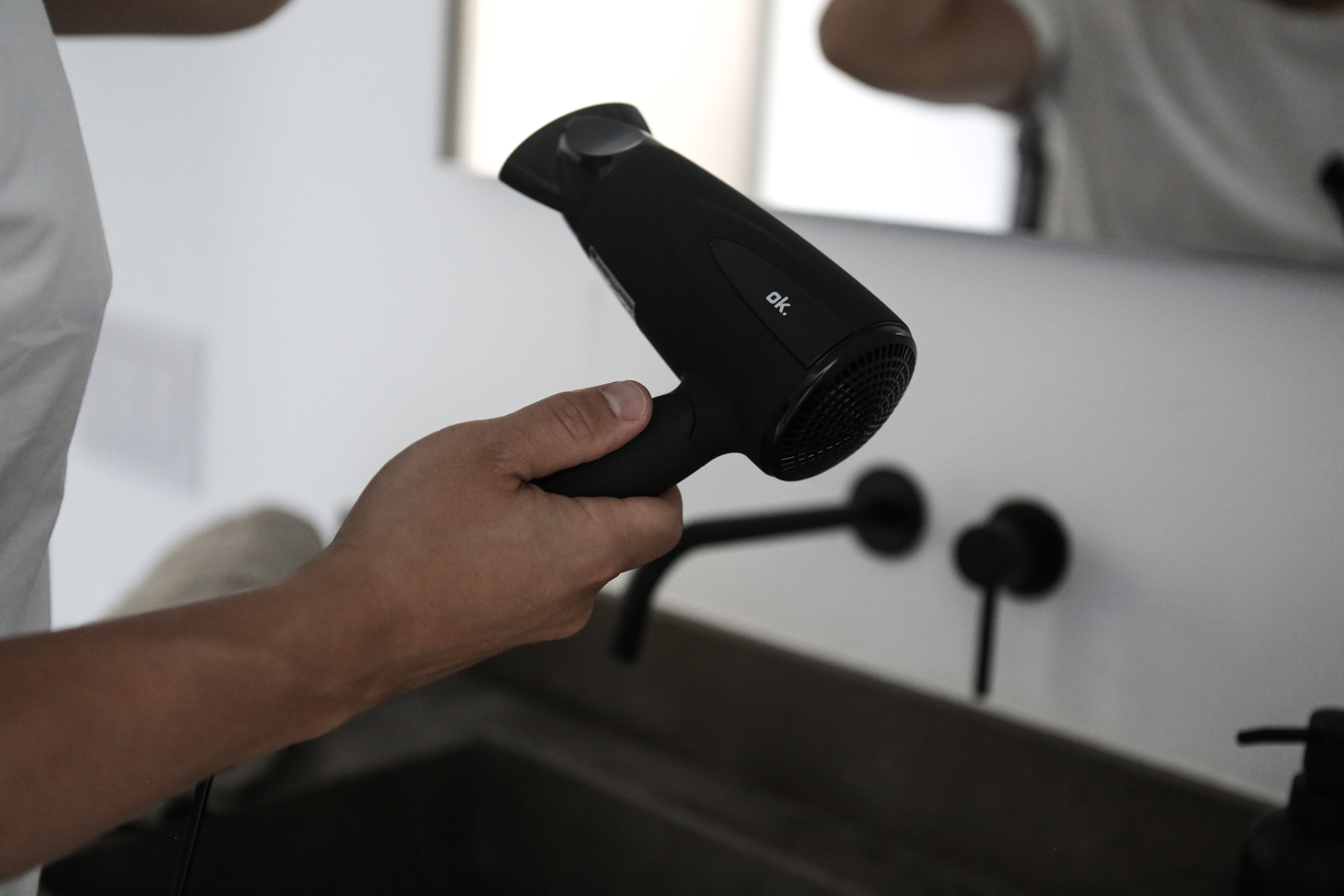 Osoba trzyma czarną suszarkę do włosów w łazience. Suszarka marki ok. Kran i lustro w tle.