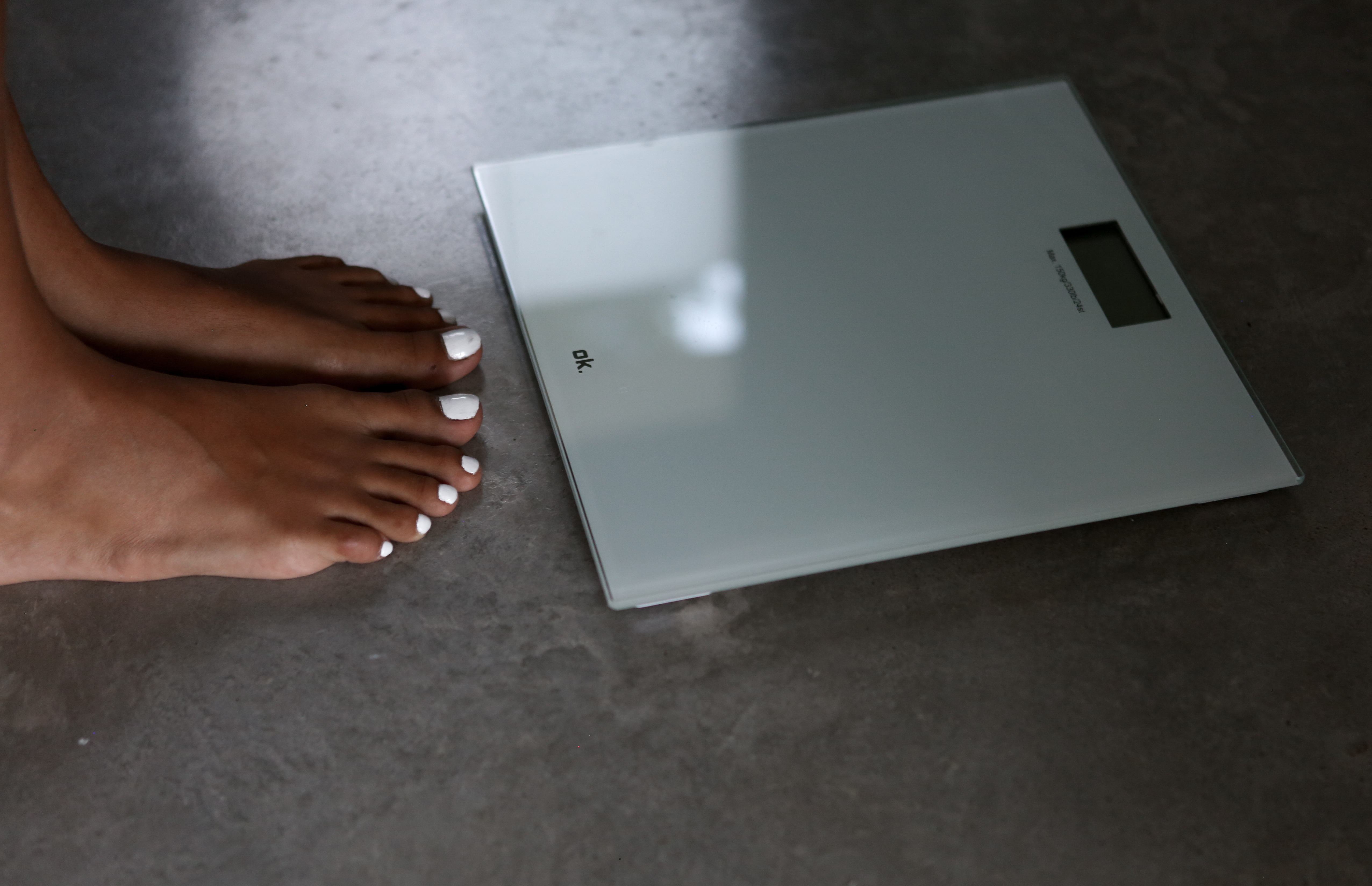 Stopy z białymi paznokciami obok białej wagi cyfrowej na szarej podłodze.