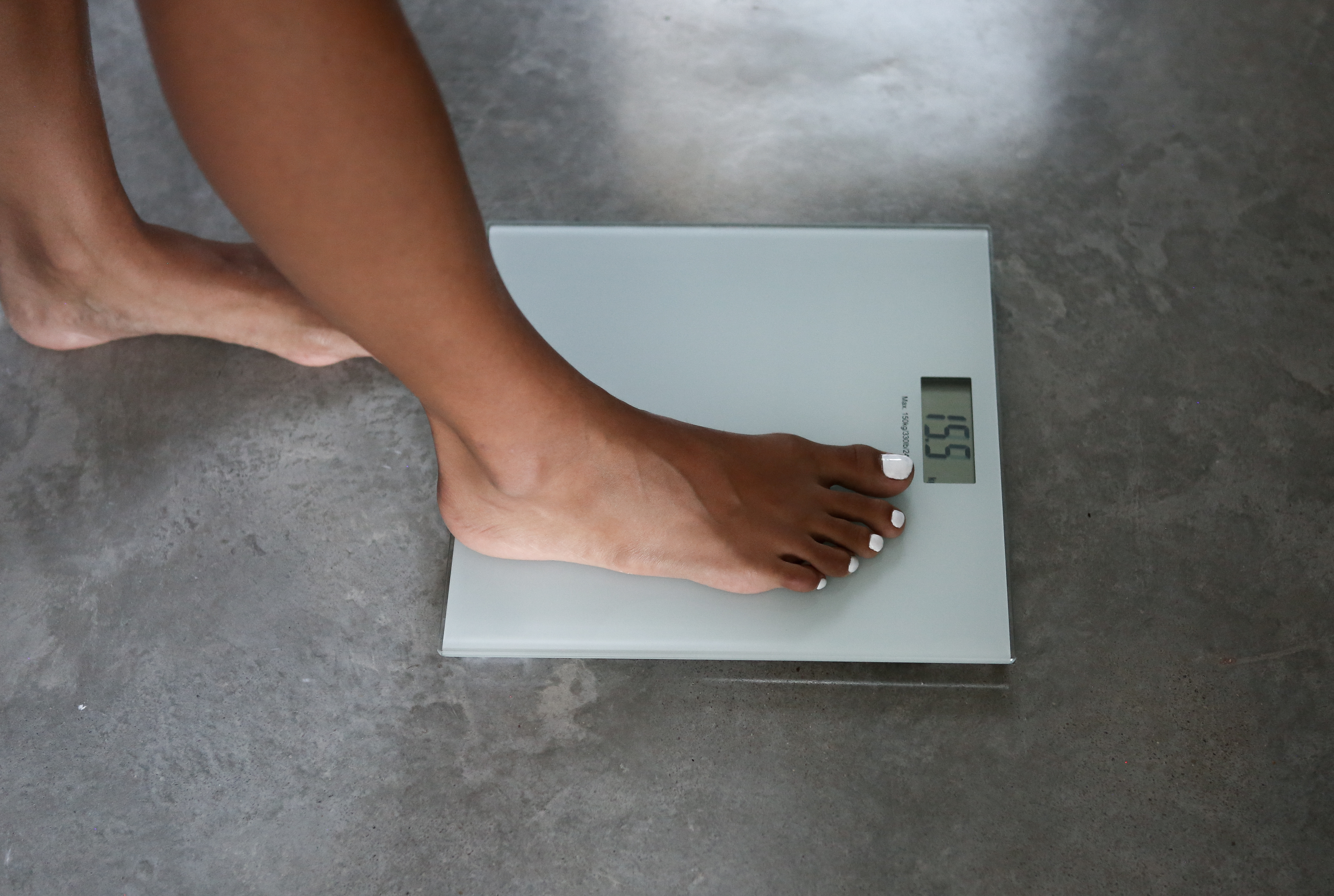 Stopa na wadze, pokazująca 155. Białe paznokcie u stóp. Ciemna skóra. Szara waga i podłoga. Mierzenie wagi.