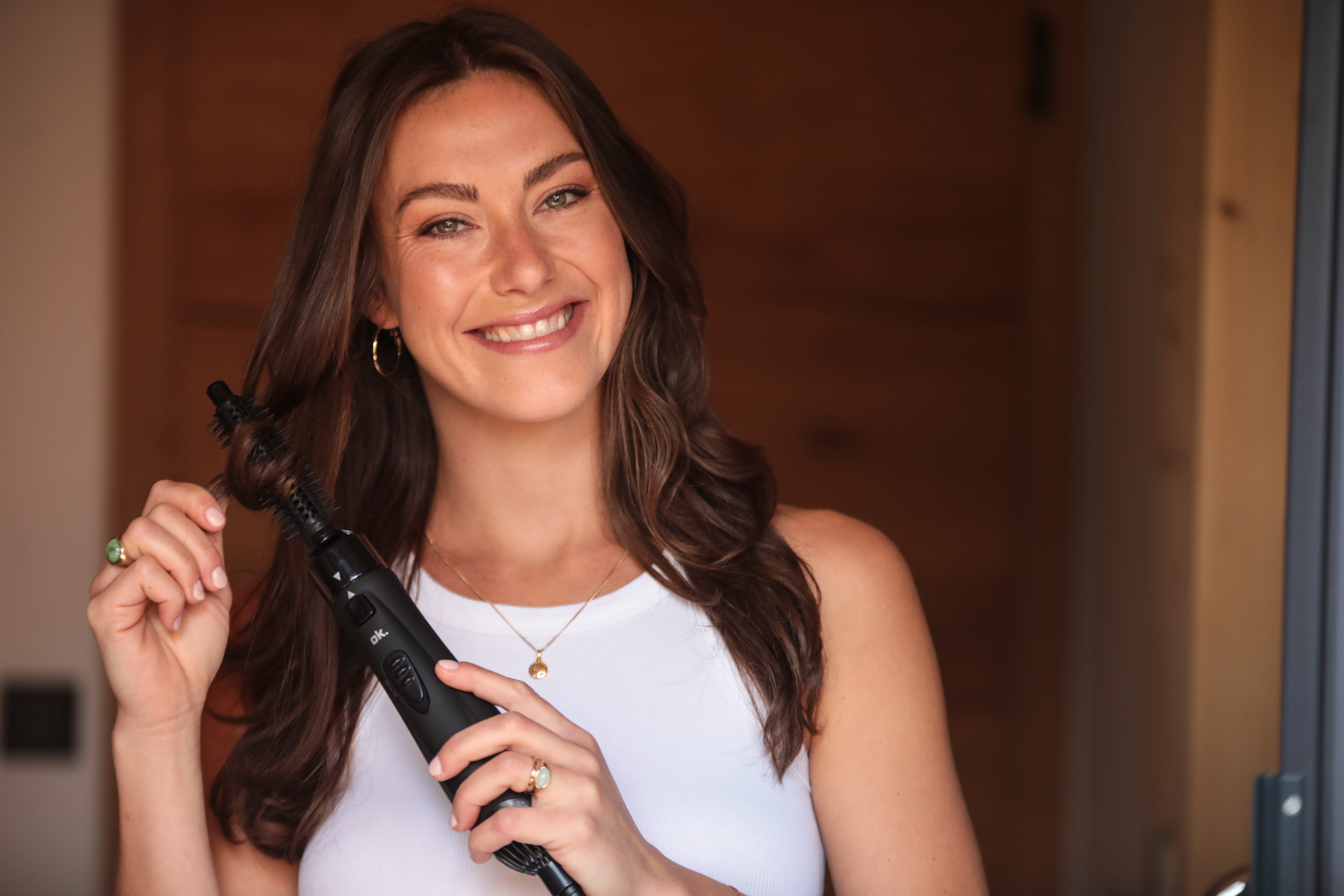 Kobieta uśmiecha się, trzymając czarne narzędzie do stylizacji włosów. Ma na sobie biały top i złotą biżuterię. Brązowe włosy i tło.
