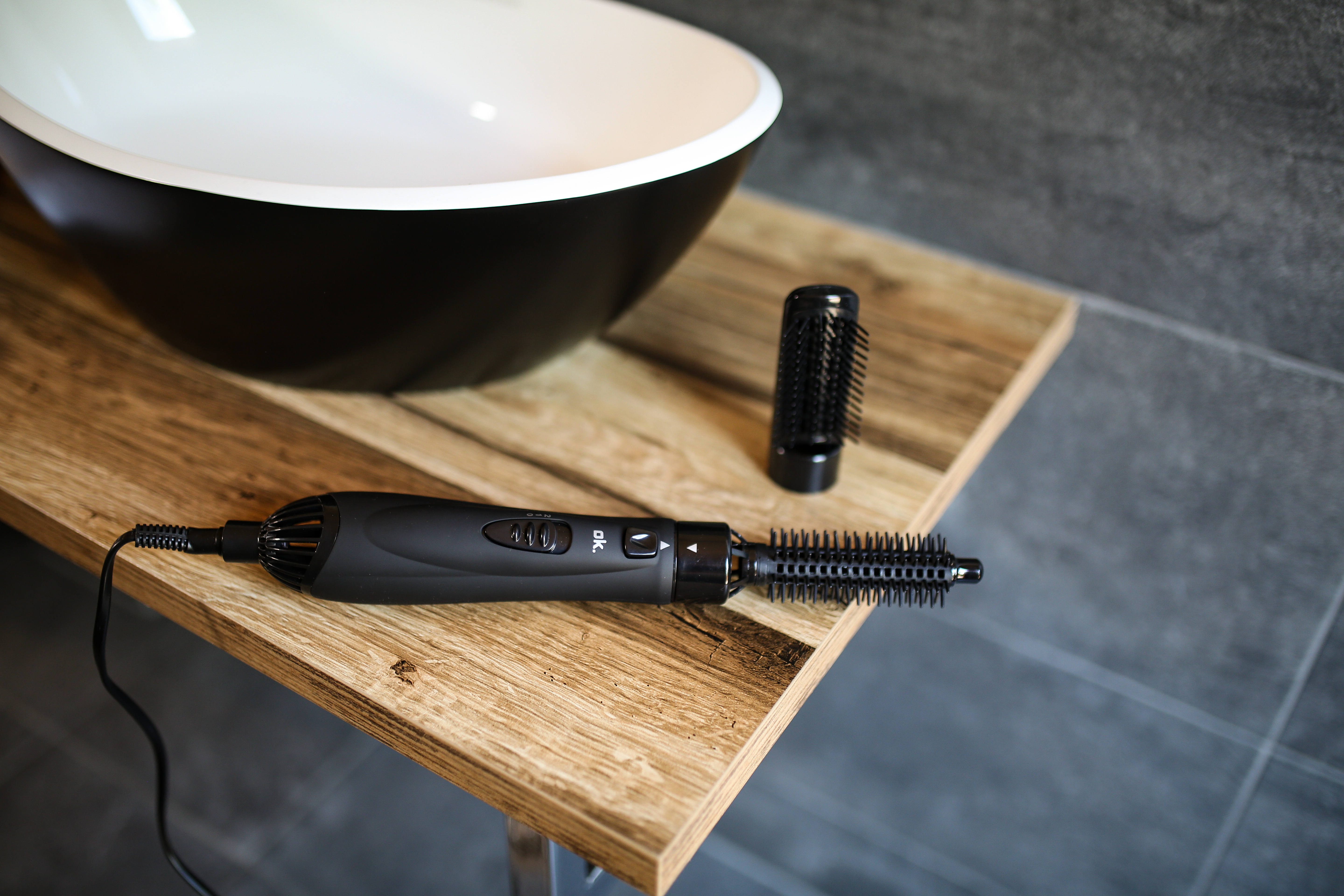 Czarny przyrząd do stylizacji włosów i szczotka spoczywają na drewnianej powierzchni. Biała i czarna umywalka w tle.