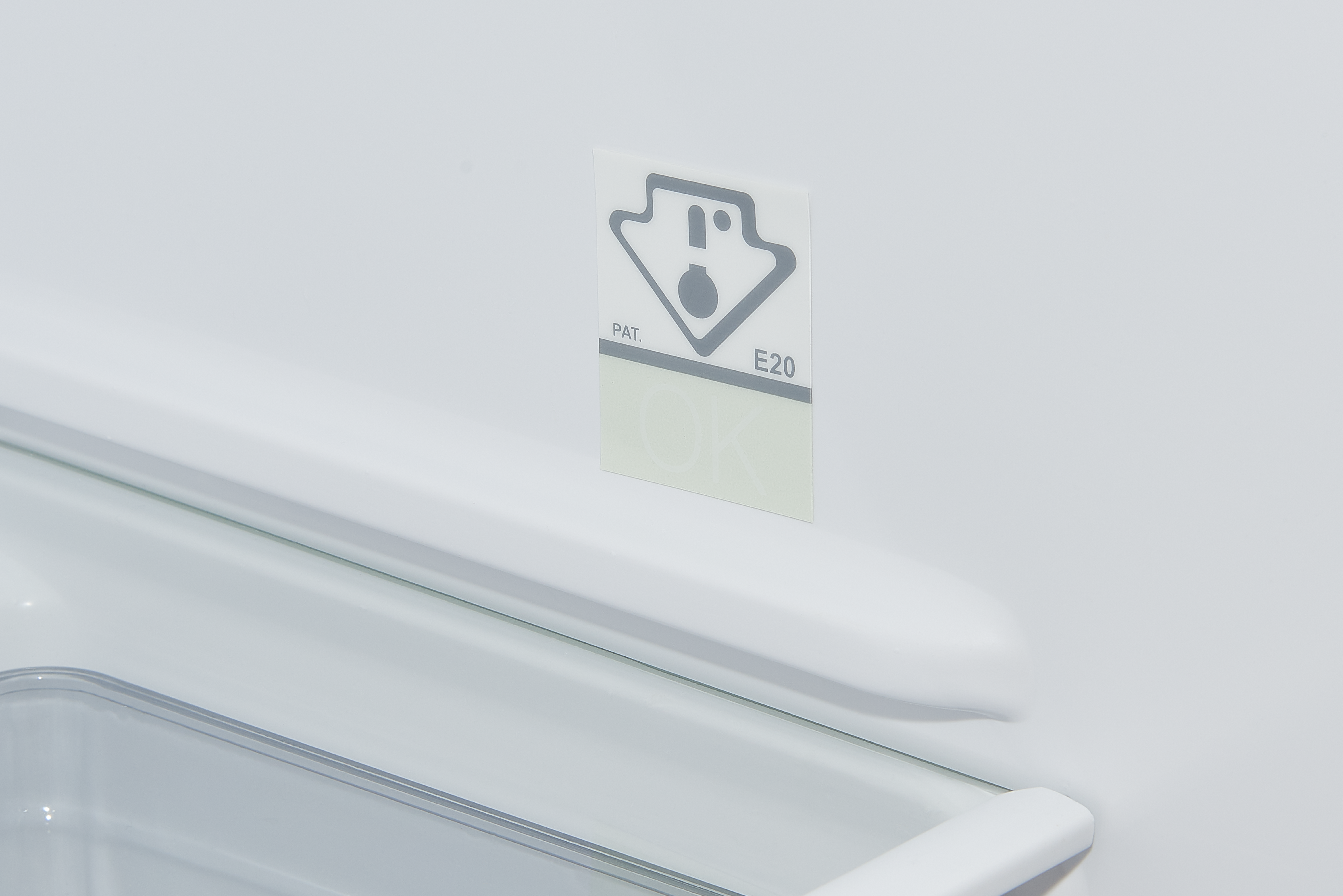 Im Inneren eines weißen Kühlschranks ist ein Aufkleber mit einem Symbol und dem Text 'PAT. E20 OK' zu sehen. Ein durchsichtiges Regal und ein Griff sind ebenfalls vorhanden.