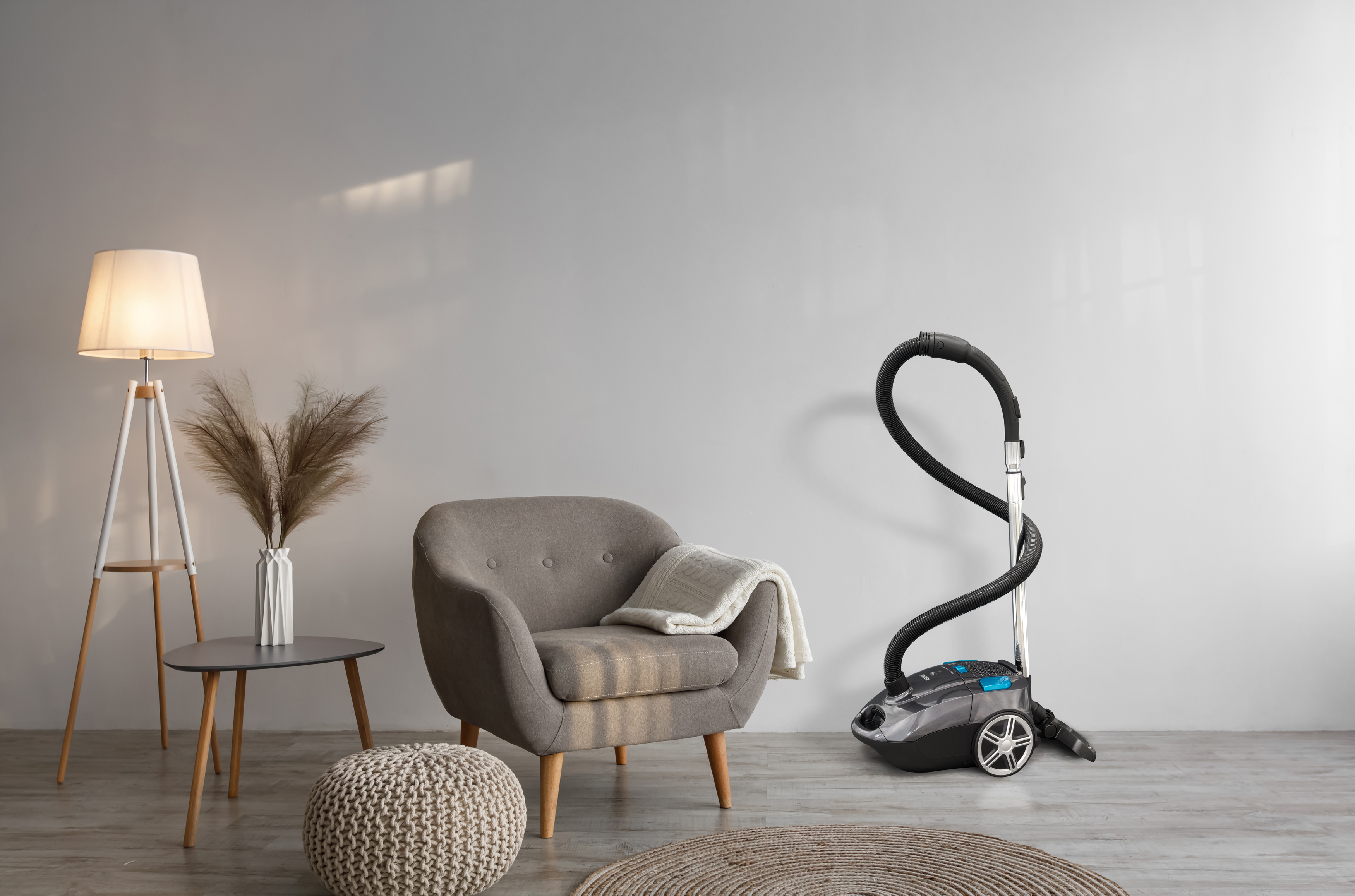 Wohnzimmerszene mit grauem Sessel, Staubsauger, Lampe und Beistelltisch. Ein Teppich und ein Sitzsack liegen auf dem Boden.