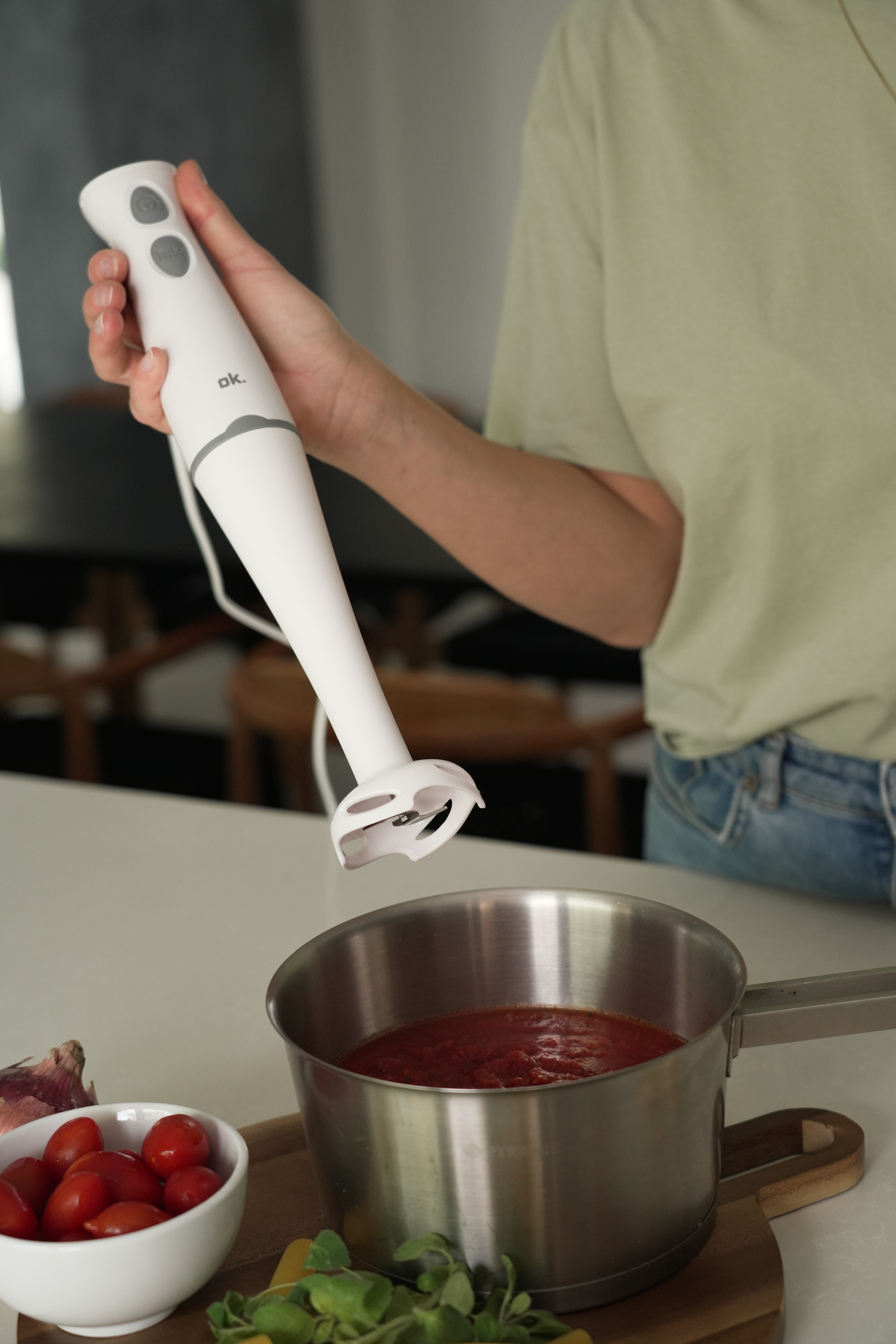 Osoba używa białego blendera ręcznego nad garnkiem z sosem pomidorowym. Składniki, takie jak pomidory i cebula, są w pobliżu.
